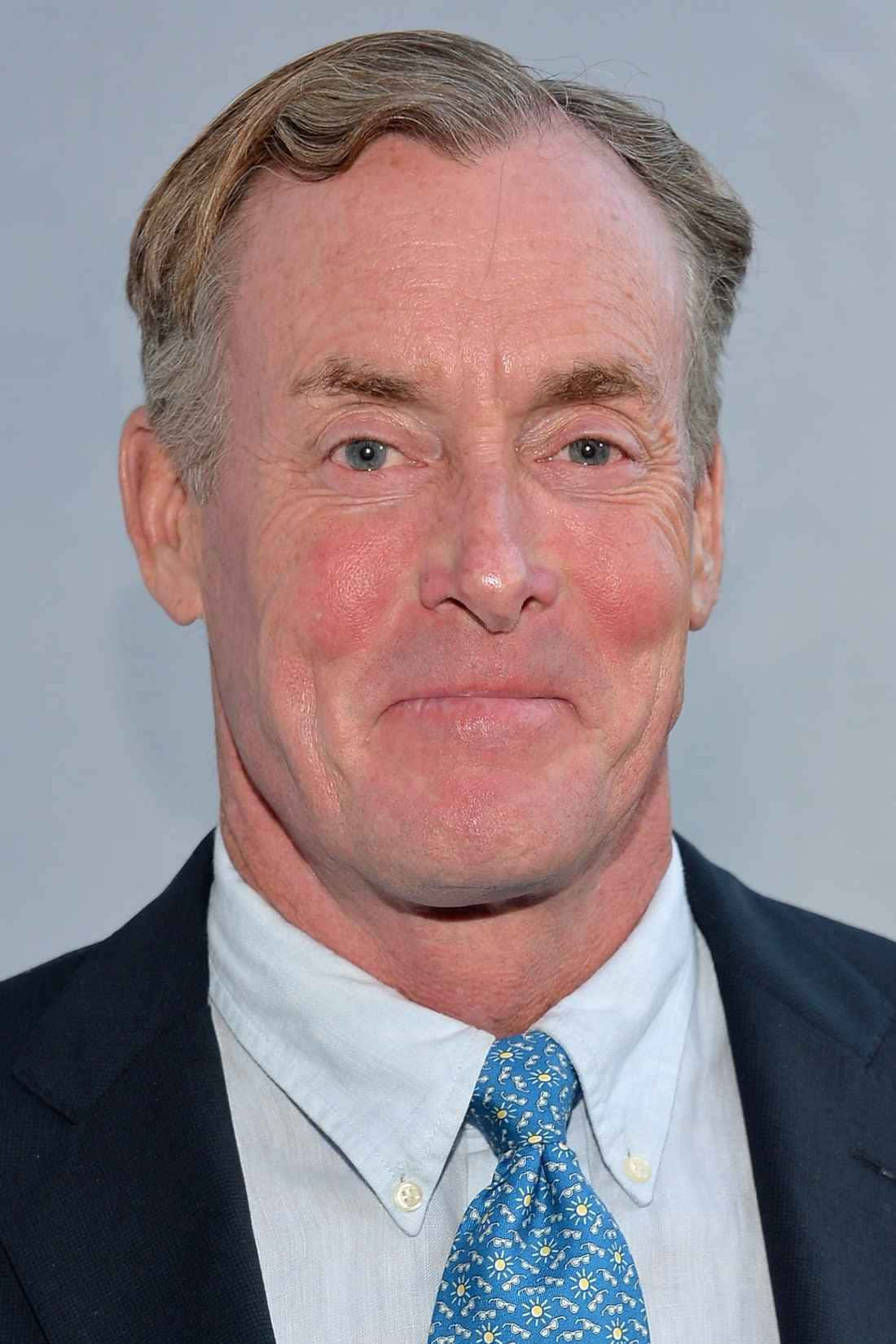 John C. Mcginley 桌布 壁紙 — 中年灰髮男士的頭像特寫，身穿深色西裝、淺藍色襯衫和圖案藍色領帶，背景為純灰色。
