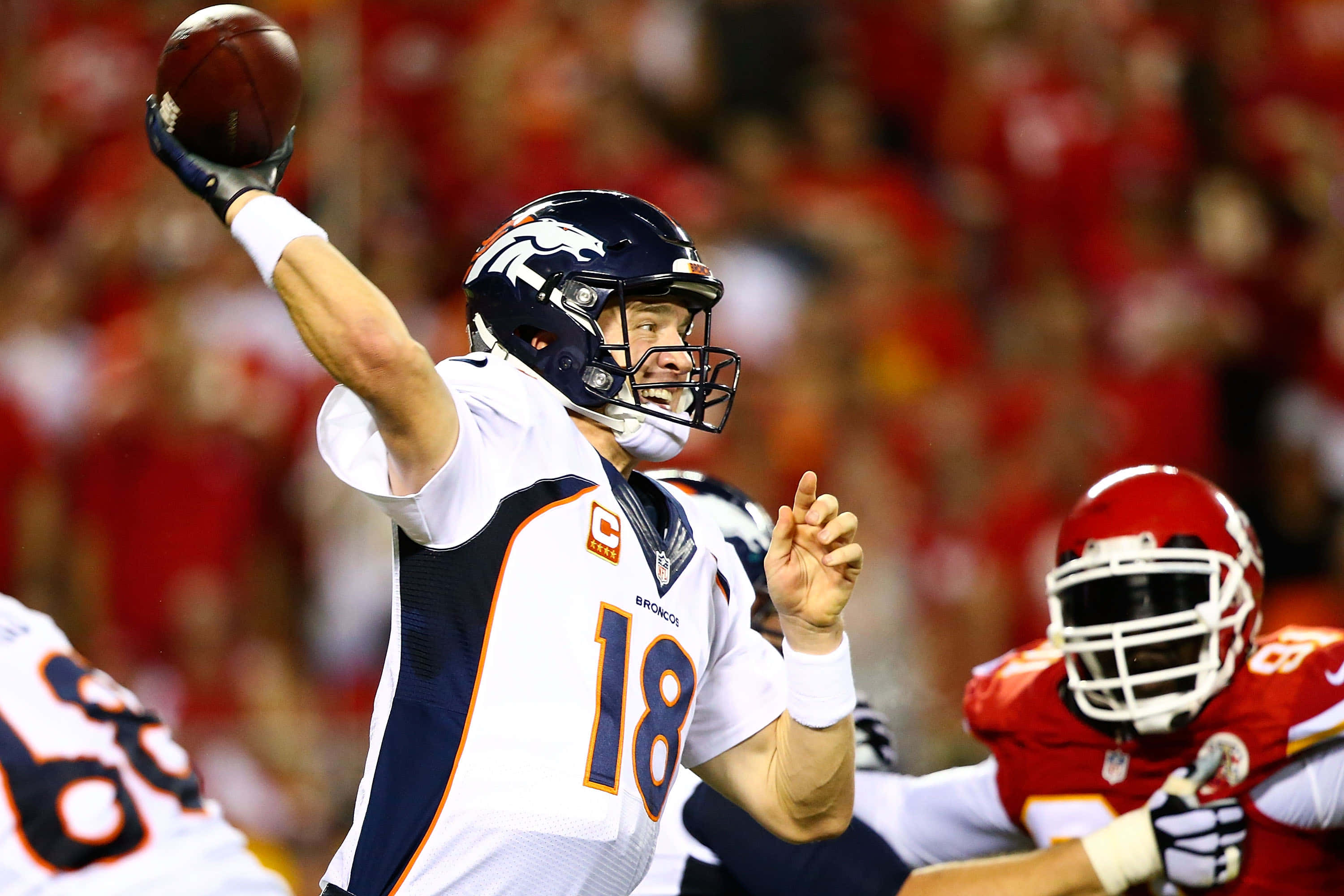 Peyton Manning 桌布 壁紙 — 丹佛野馬隊的四分衛身穿白色18號球衣，在夜間比賽中投擲橄欖球，背景是模糊的紅色體育場人群。