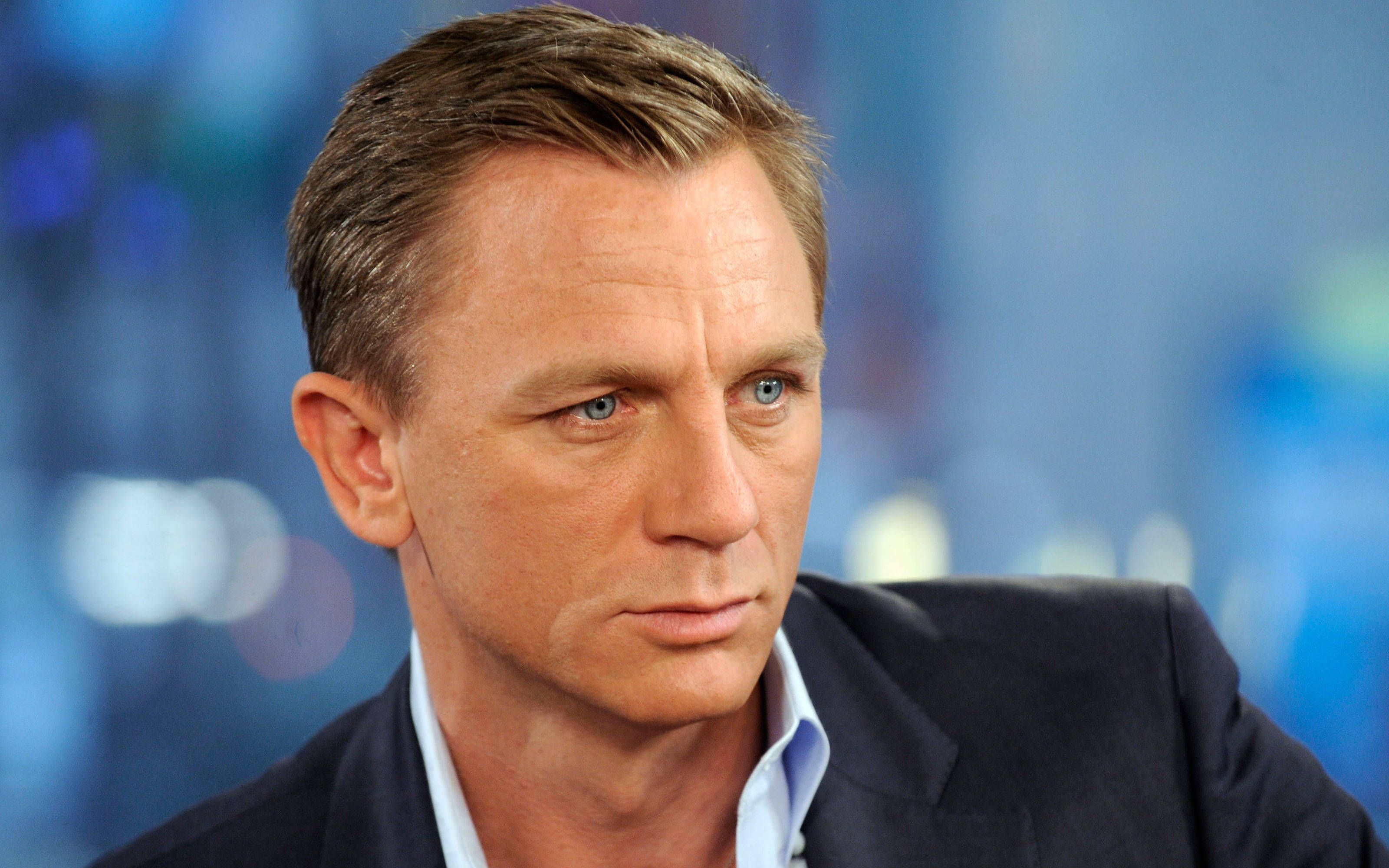 Daniel Craig 桌布 壁紙 — 丹尼爾·克雷格（Daniel Craig）的頭像特寫，擁有強烈的藍色眼睛和嚴肅的表情，背景為模糊的藍色。