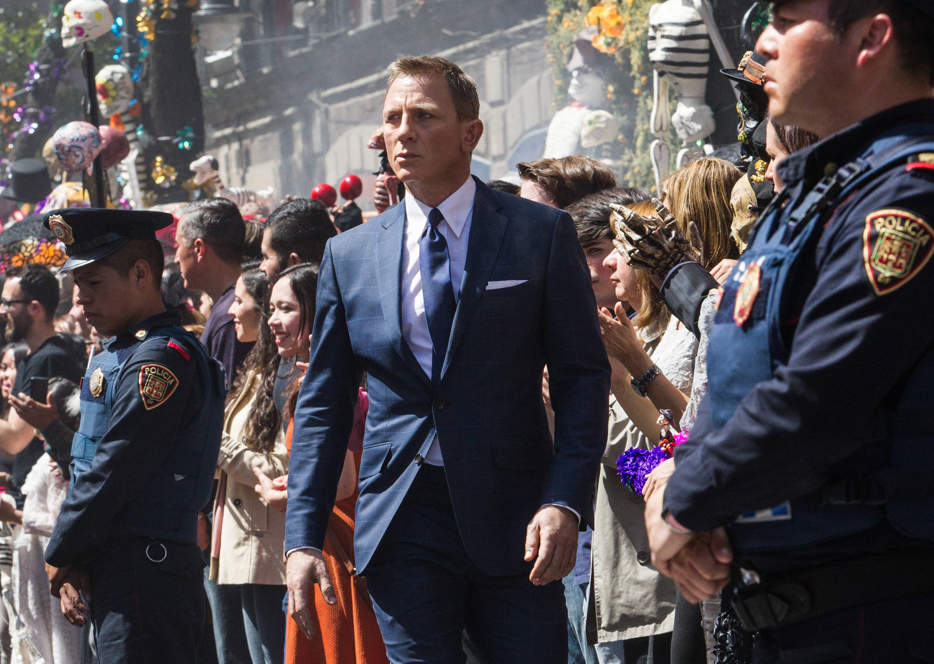 Daniel Craig 桌布 壁紙 — 丹尼爾·克雷格飾演詹姆士·龐德，自信地走過陽光普照的街頭遊行，兩旁有警察和歡呼的人群。