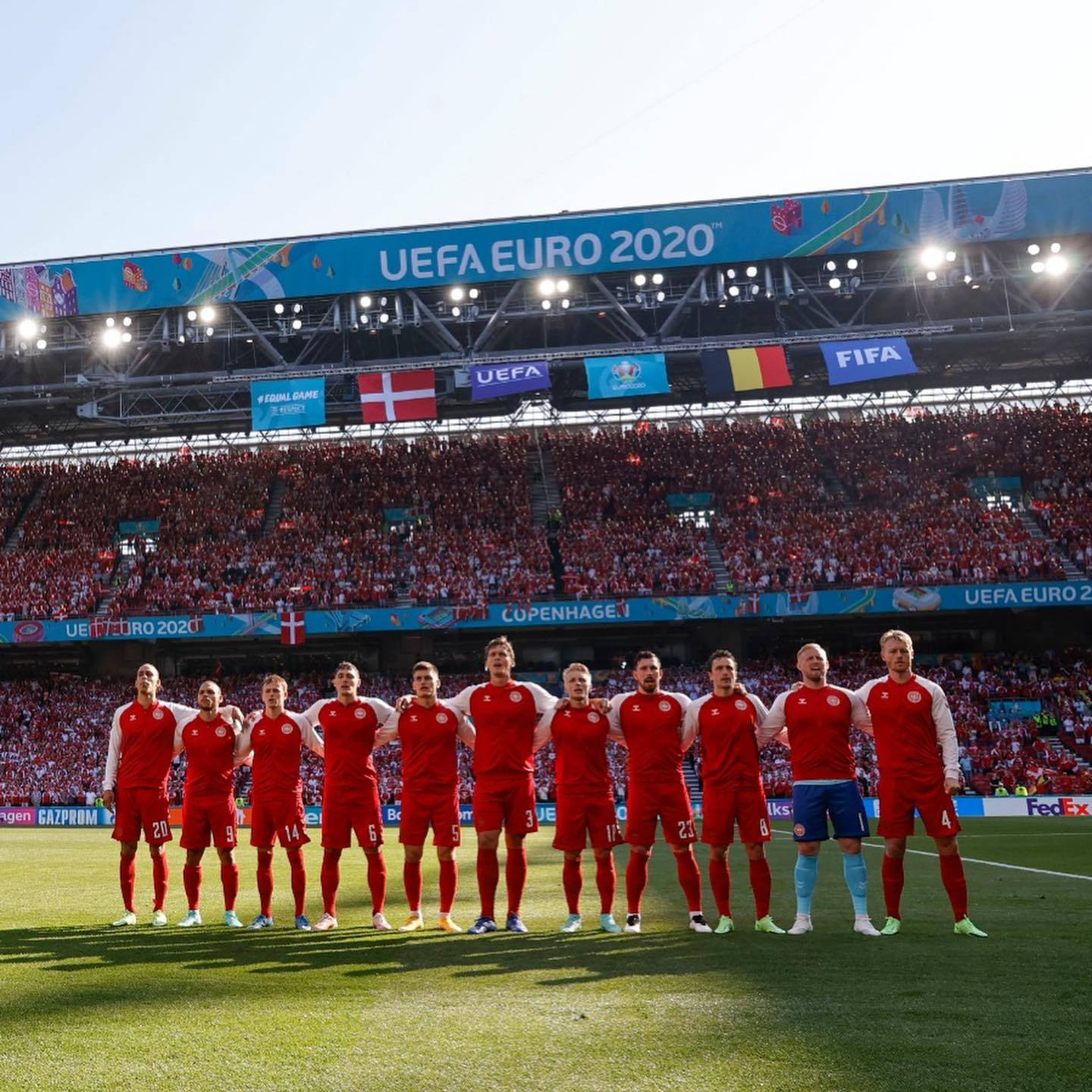 足球 桌布 壁紙 — 丹麥國家足球隊身穿紅色球衣，在 UEFA Euro 2020 的陽光普照球場上排成一列，面對座無虛席的觀眾。