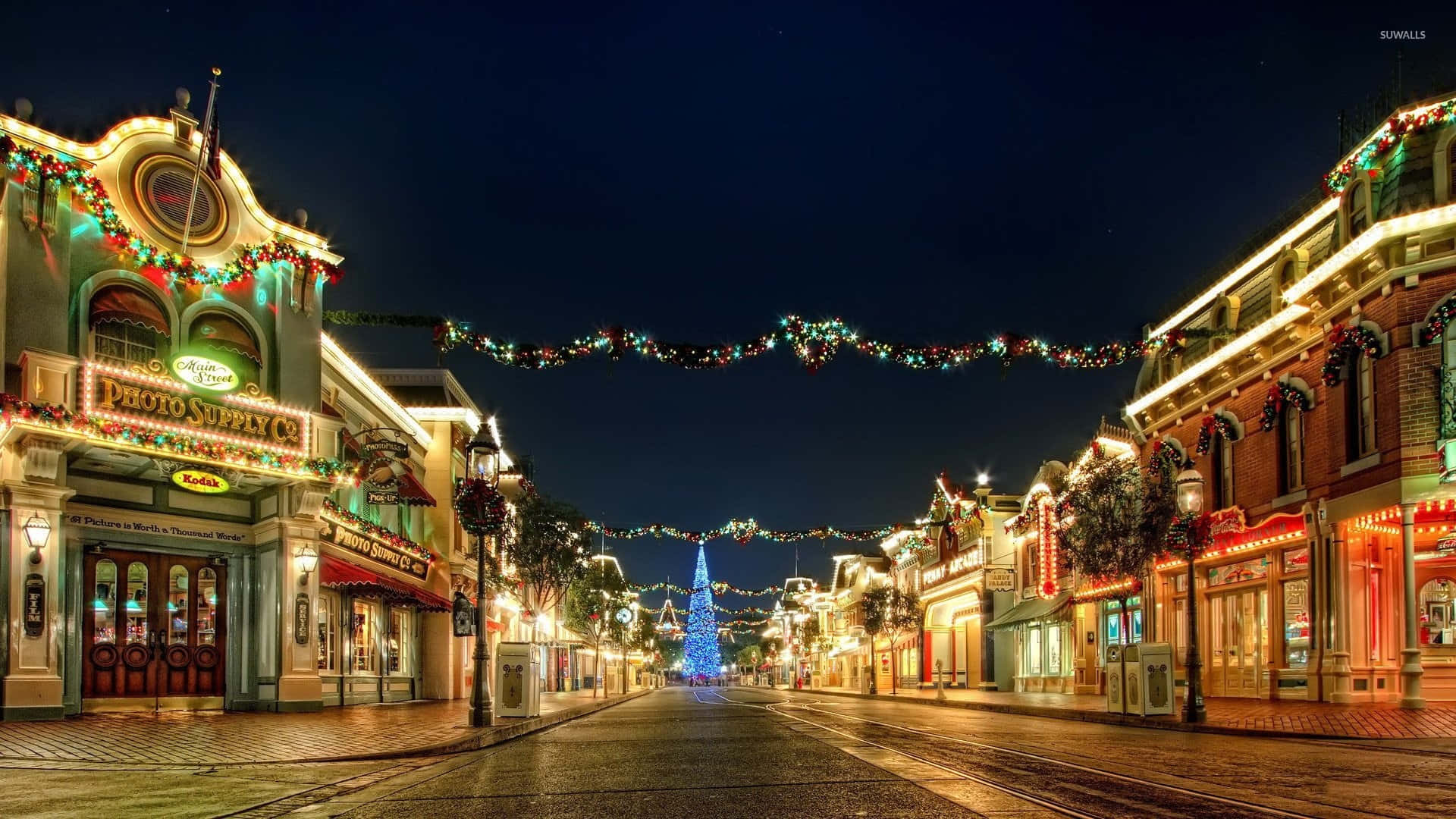 Christmas Village 桌布 壁紙 — 主題公園內一條節日氣氛濃厚的夜間街道，裝飾著發光的燈飾花環和一棵高聳的藍色聖誕樹。