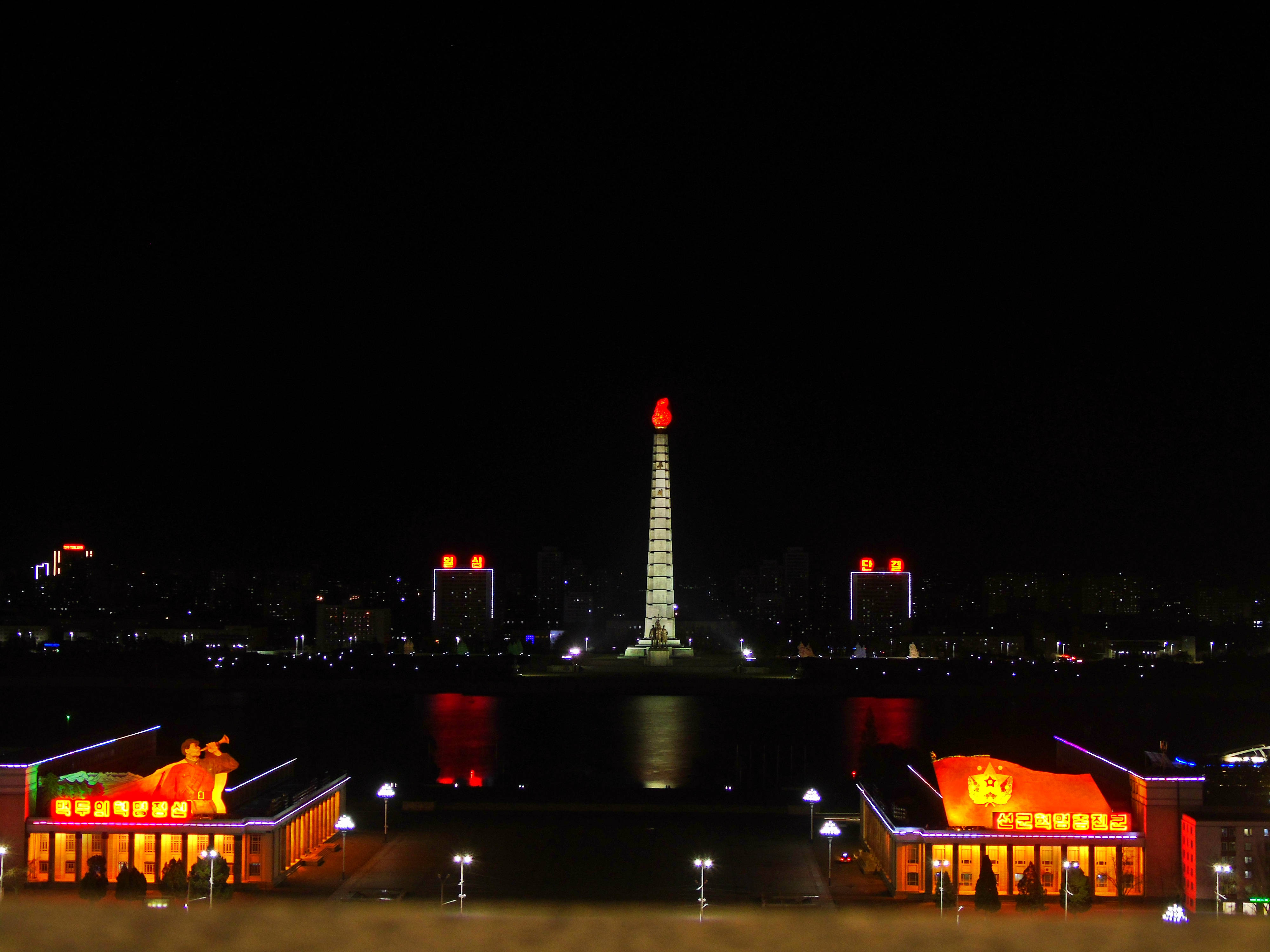 韓國 桌布 壁紙 — 主體塔在河岸上發出紅光的夜景，下方有燈火通明的建築物和反射的水面。