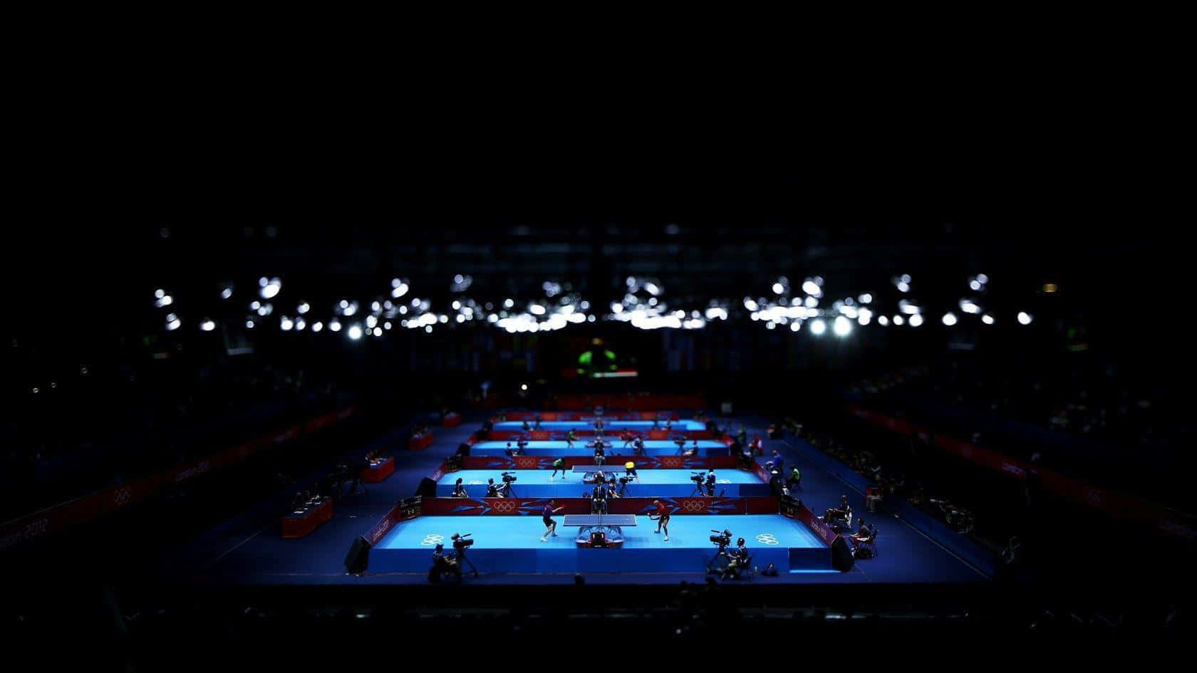 Table Tennis 桌布 壁紙 — 乒乓球運動員在黑暗場館內的藍色球場上，在明亮燈光下比賽，觀眾席模糊不清。