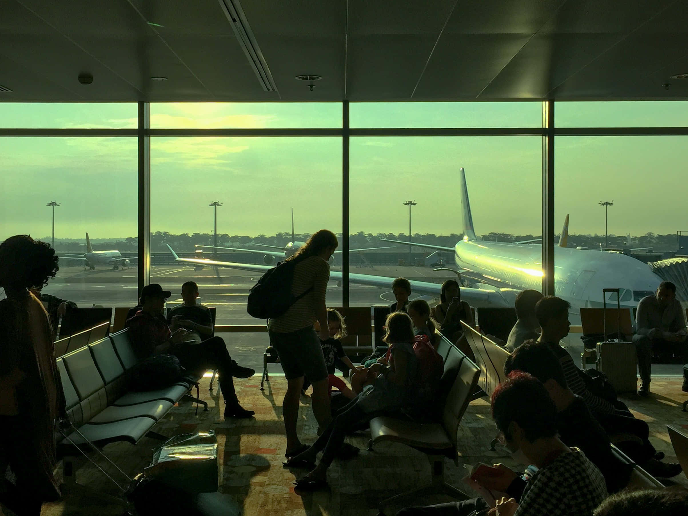Travel Desktop 桌布 壁紙 — 乘客在機場航廈內坐著和站著，觀看在金色夕陽下停機坪上的飛機。