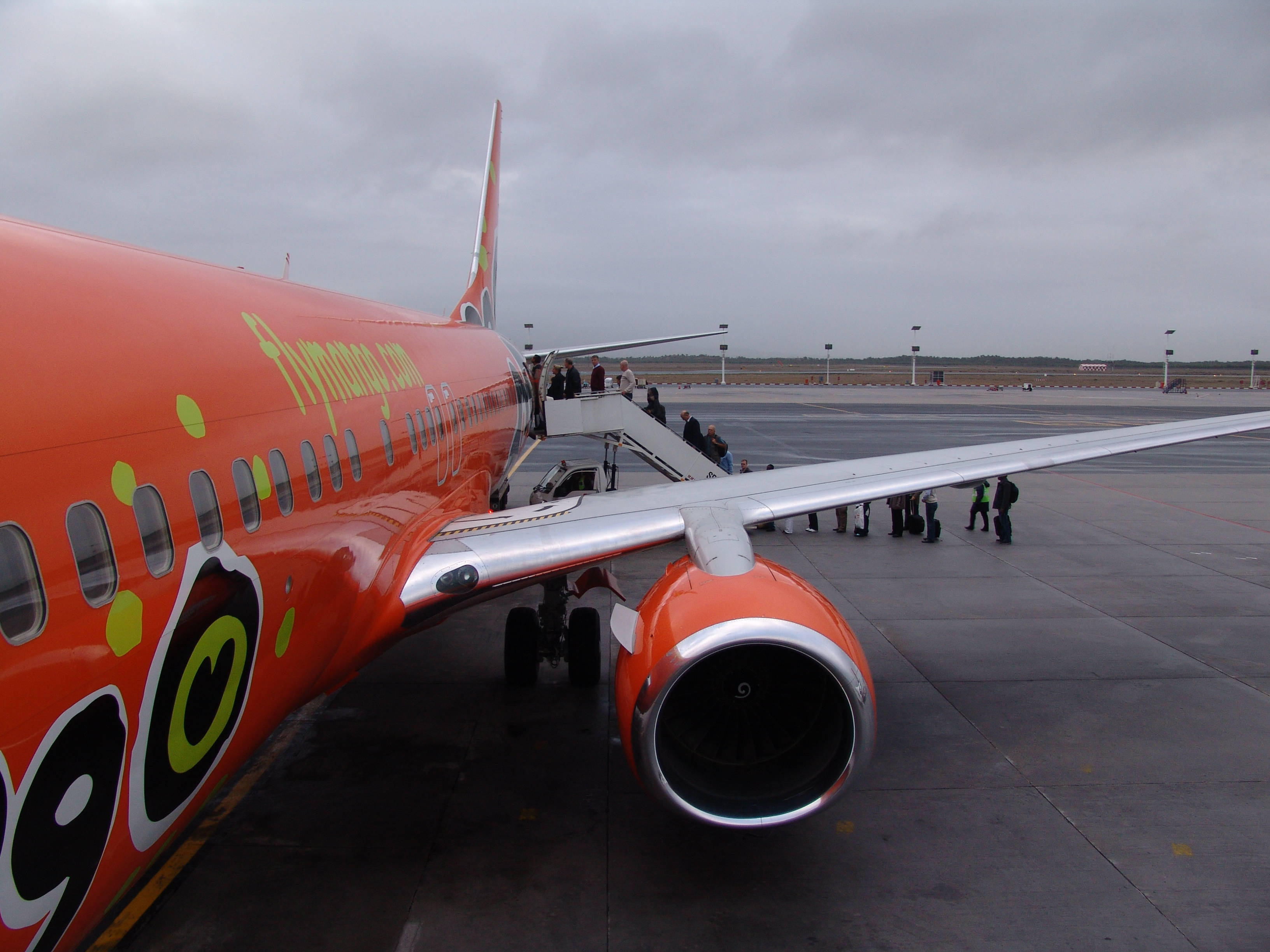 雲朵 桌布 壁紙 — 乘客在陰天的機場停機坪上，通過移動樓梯登上明亮的橙色廉價航空公司噴射機。