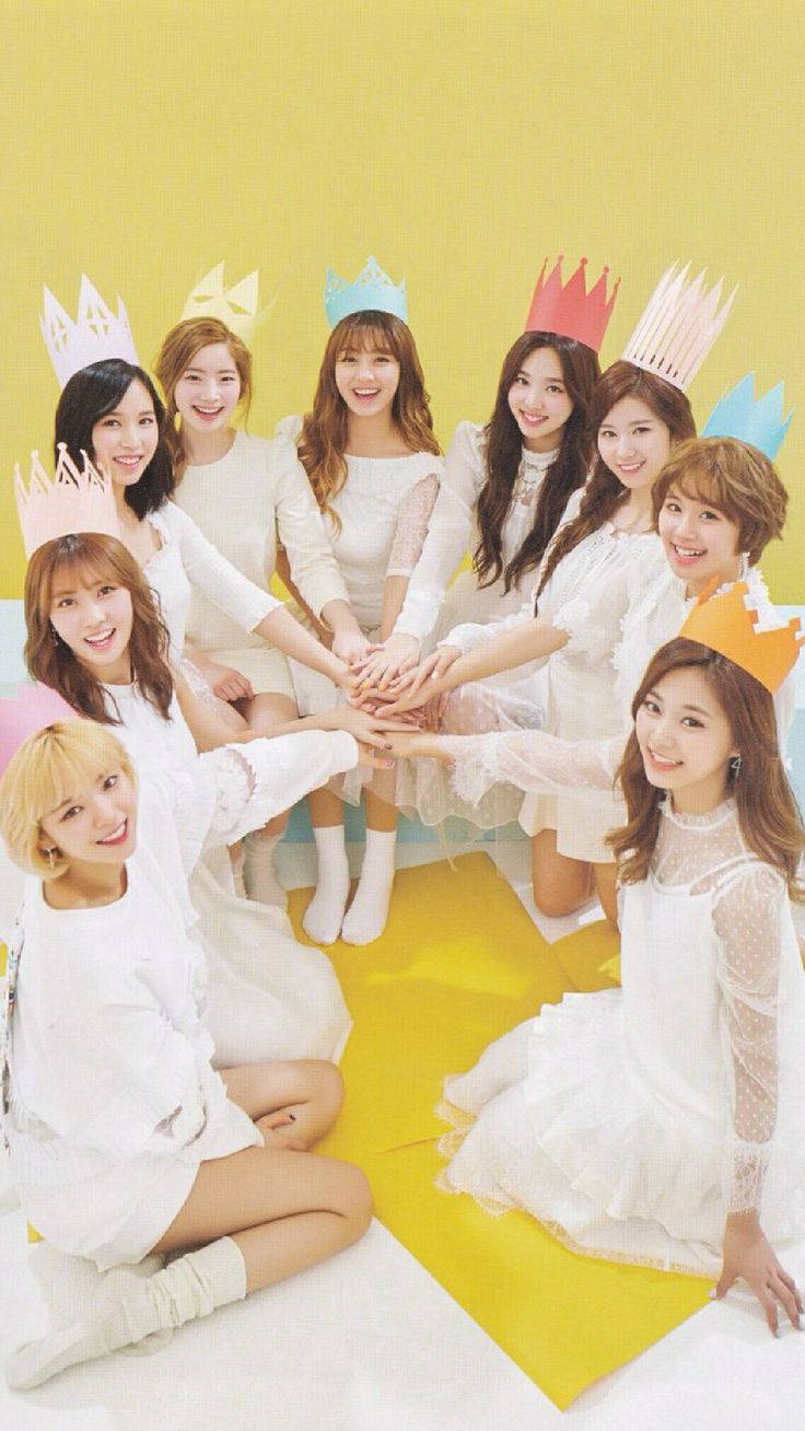 twice 桌布 壁紙 — 九位微笑的女性穿著白色連衣裙和彩色紙冠，在明亮的黃色背景前圍成一個圓圈疊手。