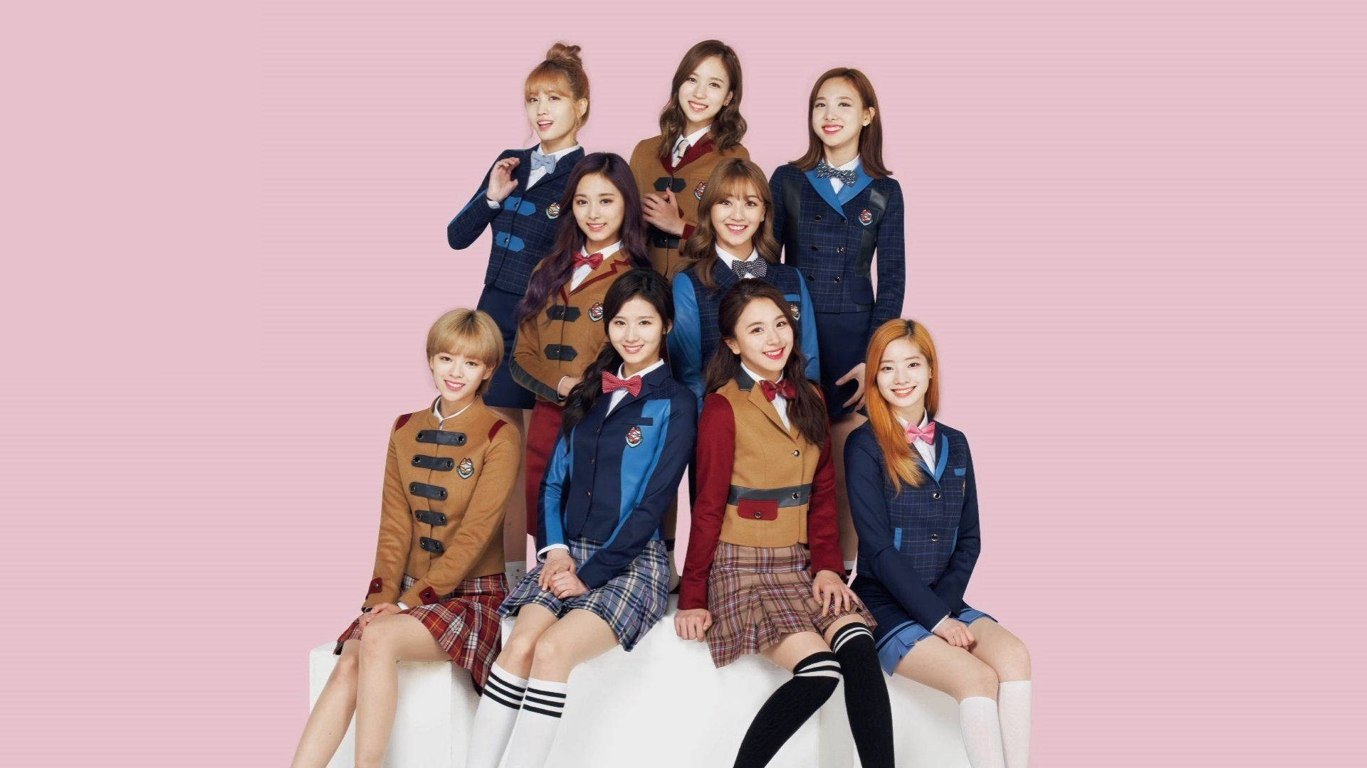 twice 桌布 壁紙 — 九位微笑的年輕女性穿著協調的校服，在純粉紅色背景前開心地擺姿勢。