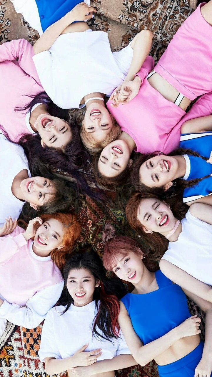 twice 桌布 壁紙 — 九位微笑的年輕女性穿著粉紅色和藍色的服裝，躺在圖案地毯上圍成一個圓圈，快樂地向上看。