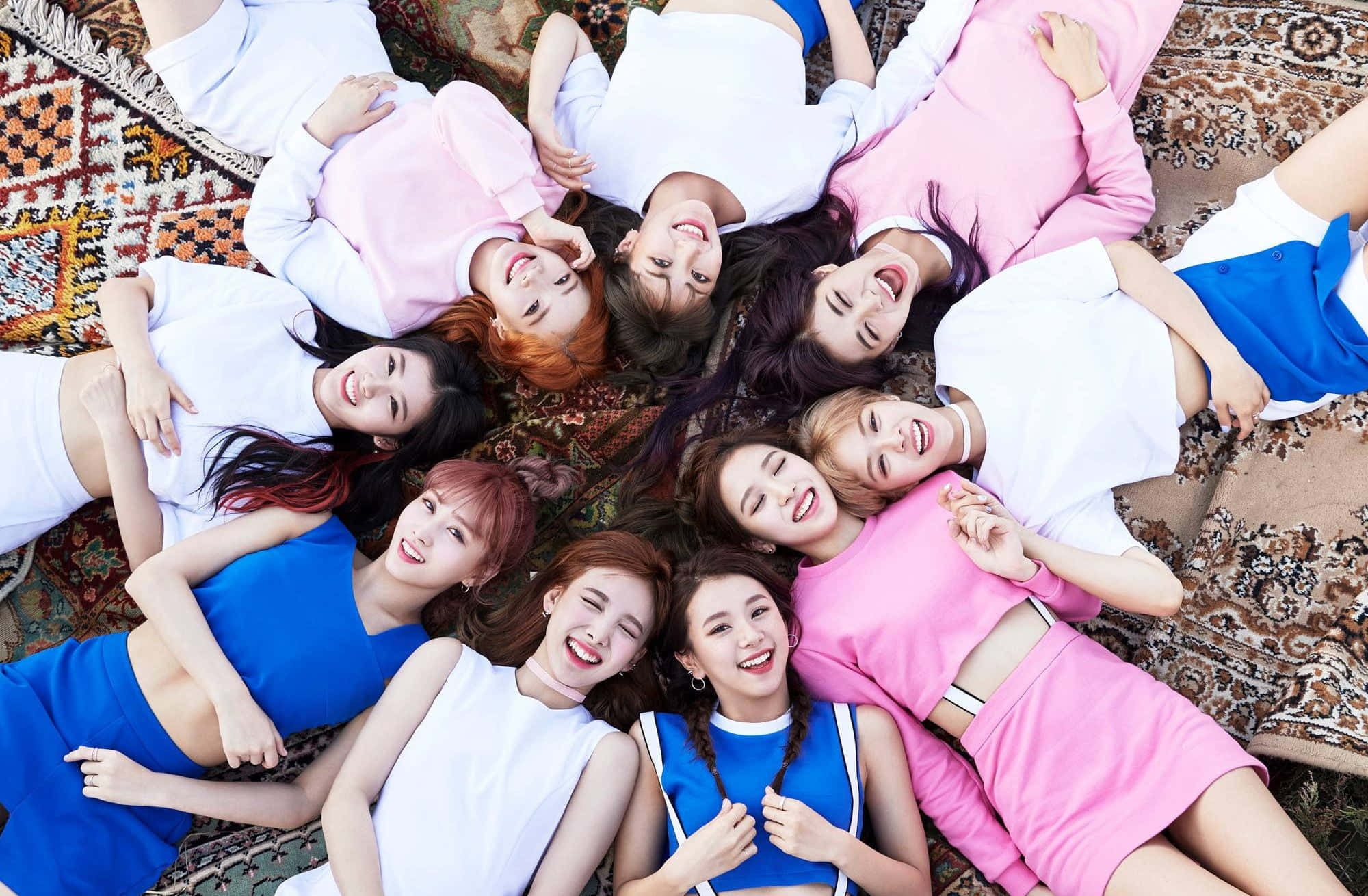 twice 桌布 壁紙 — 九位微笑的年輕女性穿著色彩繽紛的服裝，躺在圖案地毯上圍成一個圓圈，抬頭看著相機。