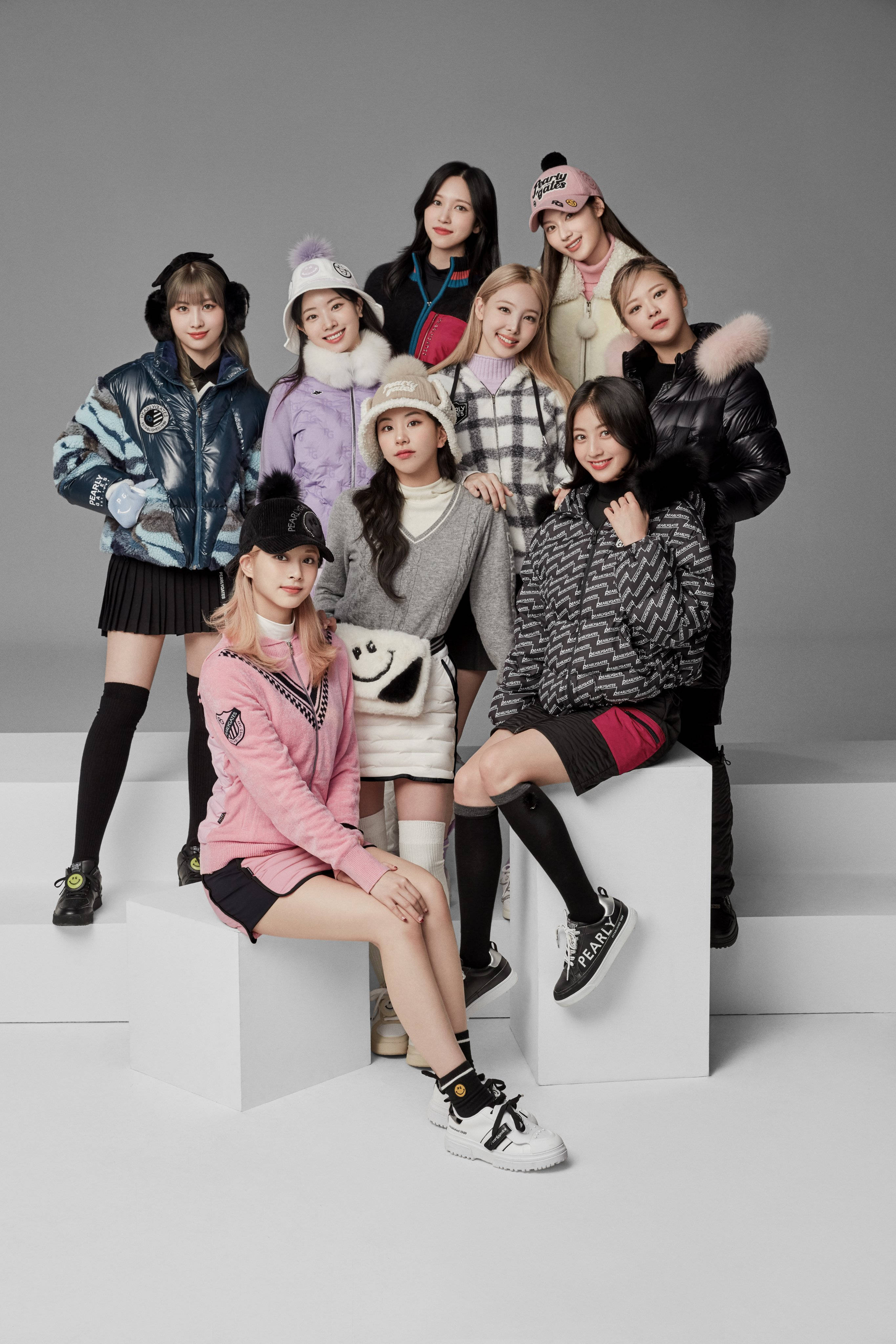 Kpop Aesthetic 桌布 壁紙 — 九位時尚年輕女性穿著潮流冬季外套和配飾，在白色方塊上自信擺姿勢，背景為中性灰色攝影棚。
