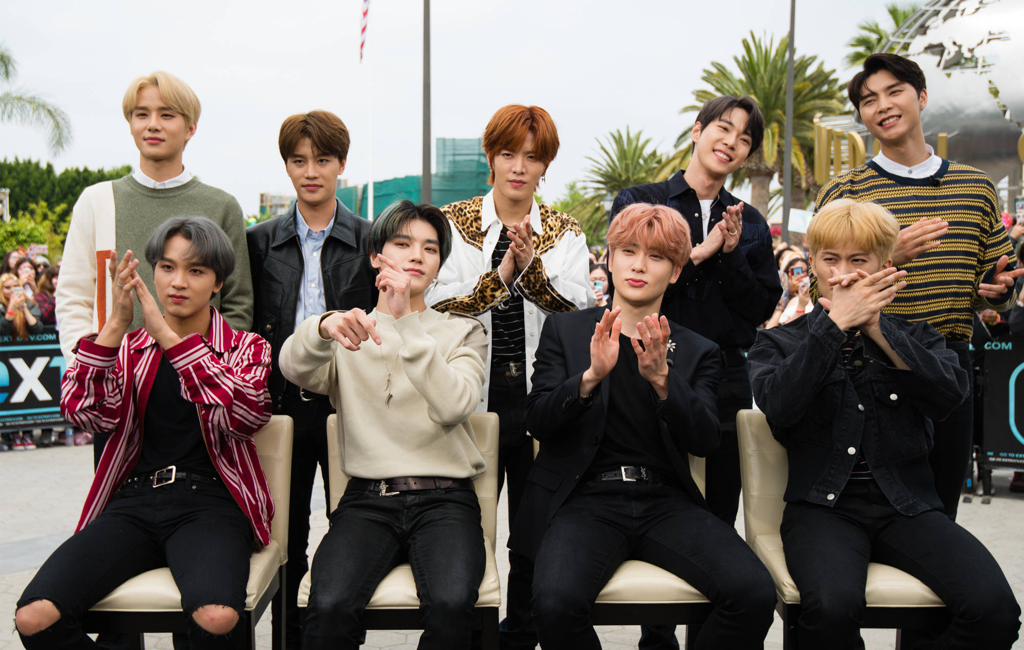 Nct 桌布 壁紙 — 九位時尚年輕男士穿著潮流服裝，在戶外與棕櫚樹和身後的人群一起愉快地擺姿勢。