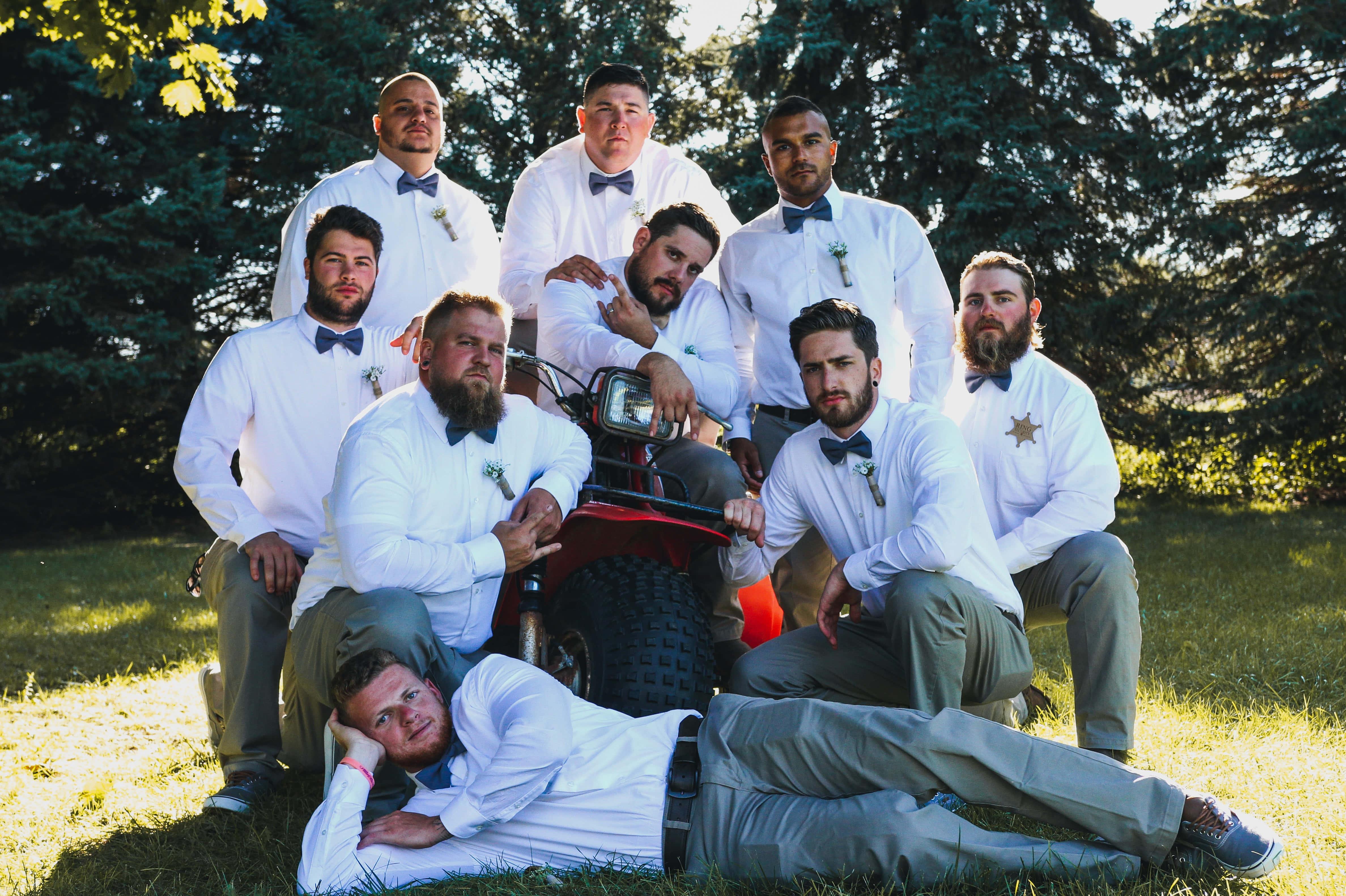 Groomsman 桌布 壁紙 — 九位穿著白襯衫和領結的伴郎在陽光明媚的草地上，身後有樹木，圍著一輛紅色ATV頑皮地擺姿勢。
