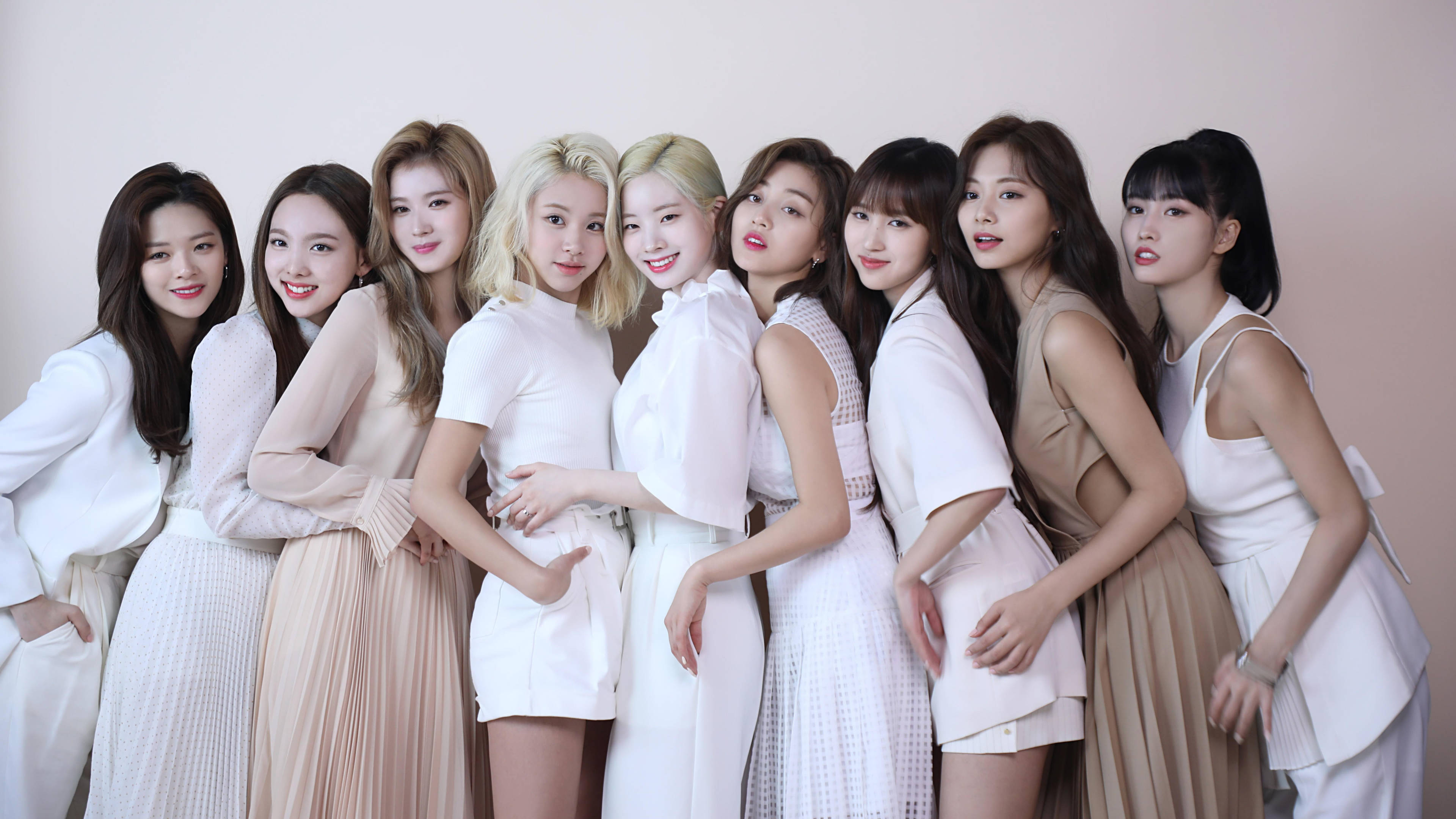 Kpop Aesthetic 桌布 壁紙 — 九位身穿白色和米色服裝的女性在柔和粉紅色背景和溫暖燈光下緊密地擺姿勢。