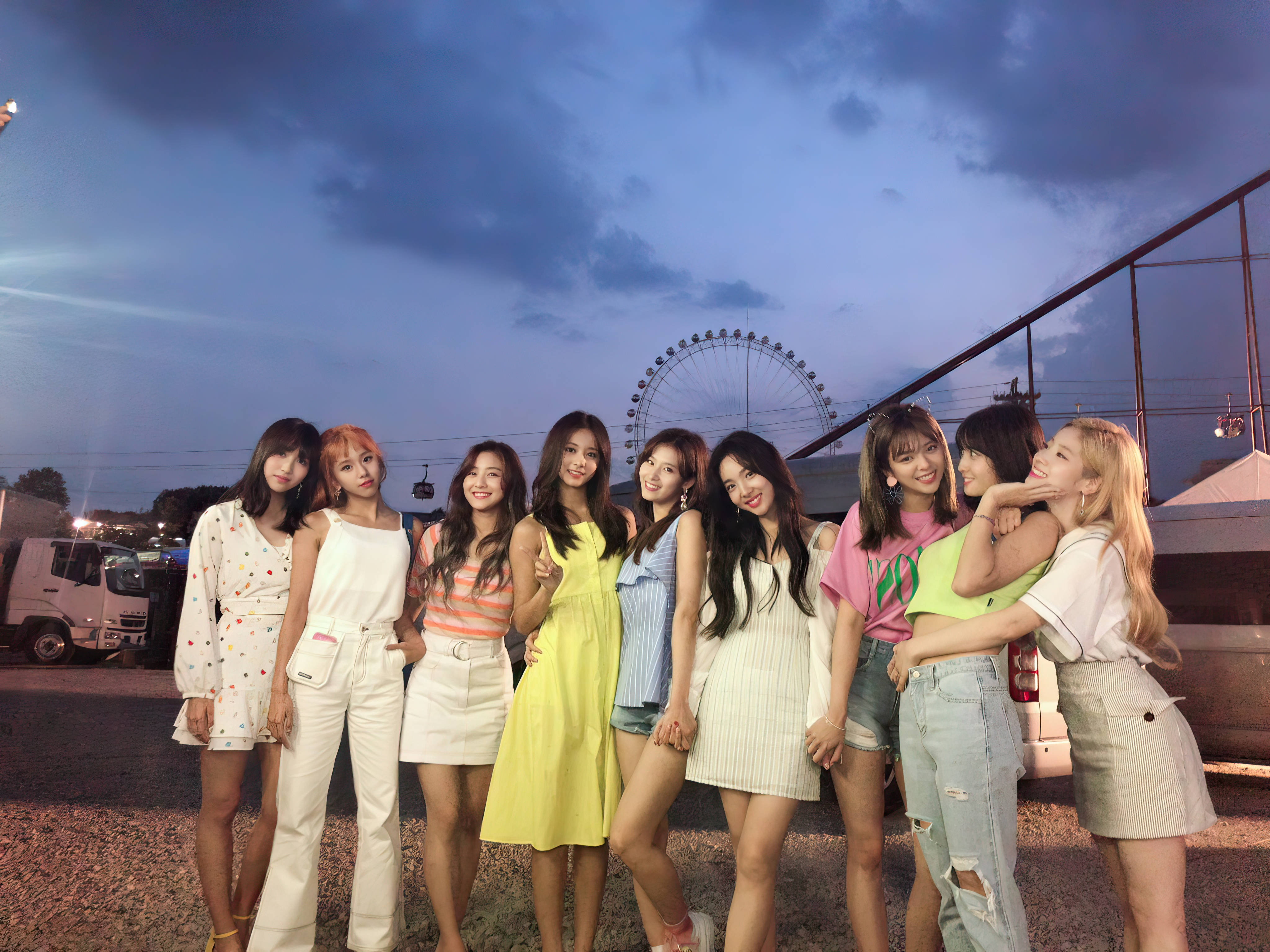 Kpop Aesthetic 桌布 壁紙 — 九位身穿色彩繽紛夏日服裝的年輕女性，在黃昏時分於摩天輪附近，在多雲的天空下愉快地擺姿勢。
