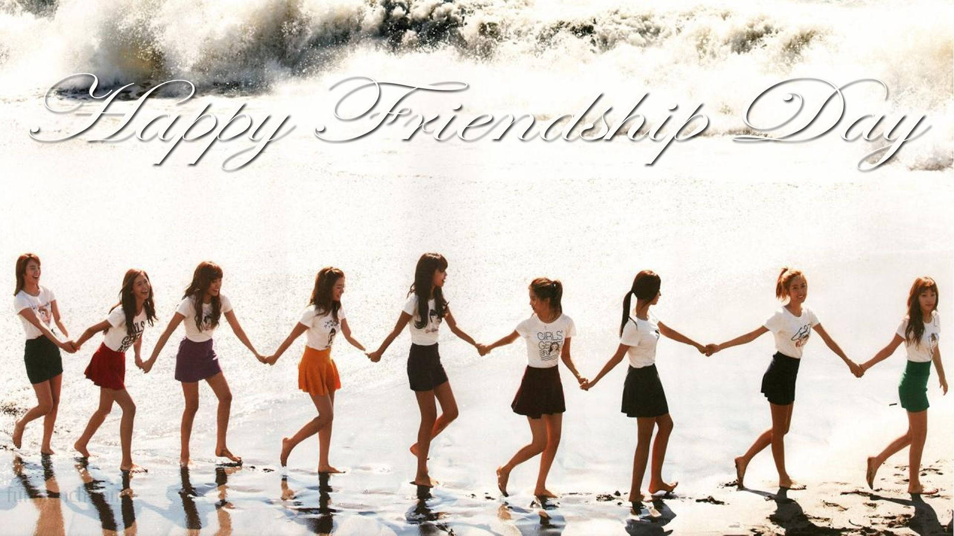 Friendship Day 桌布 壁紙 — 九個穿著白襯衫和彩色裙子的女孩手牽手沿著陽光明媚的海灘行走，身後有拍岸的海浪。