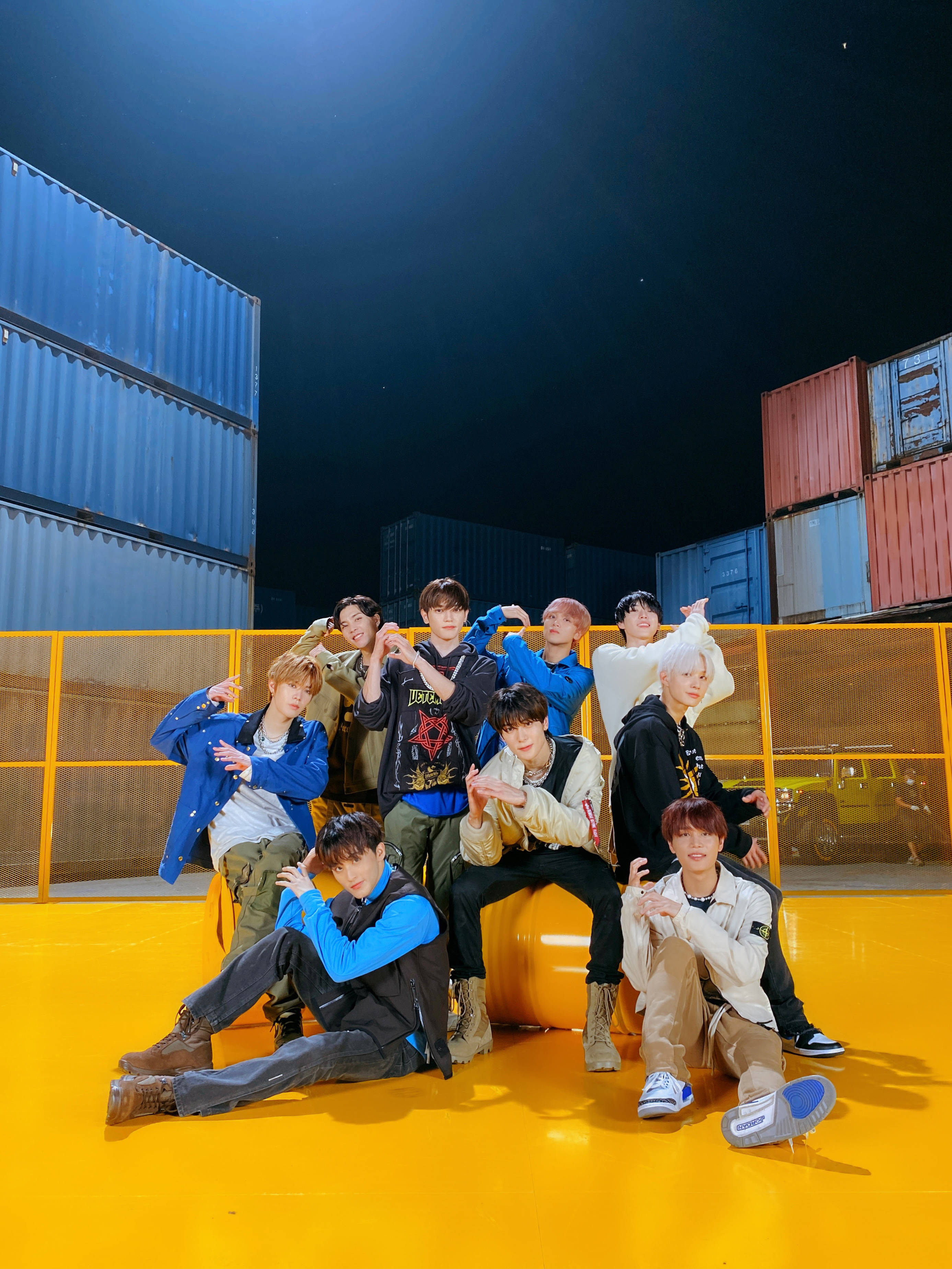 Nct 桌布 壁紙 — 九名年輕男子在明亮的黃色地板上自信地擺姿勢，背景是堆疊的貨運集裝箱，上方是繁星點點的夜空。