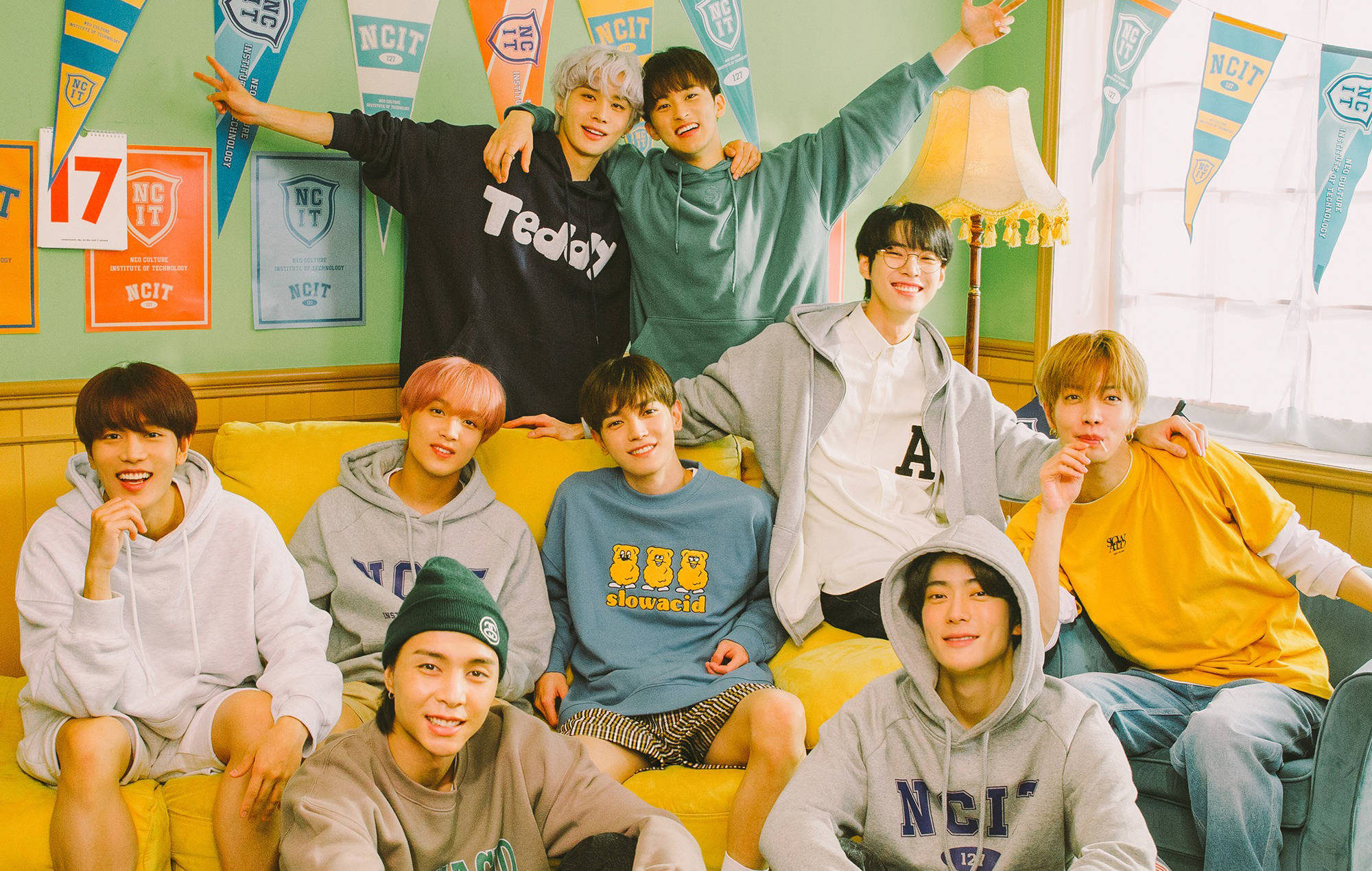 Nct 桌布 壁紙 — 九名年輕男子穿著休閒連帽衫和T恤，在裝飾有NCIT橫幅的明亮房間裡的黃色沙發上愉快地擺姿勢。