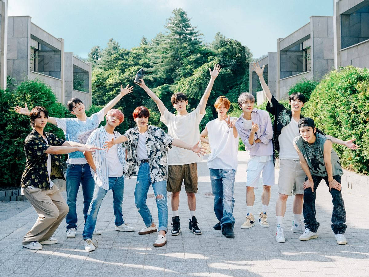 Nct 桌布 壁紙 — 九名年輕男子身穿休閒服裝，在陽光明媚的鋪砌庭院中擺出動態姿勢，周圍環繞著綠化和現代建築。