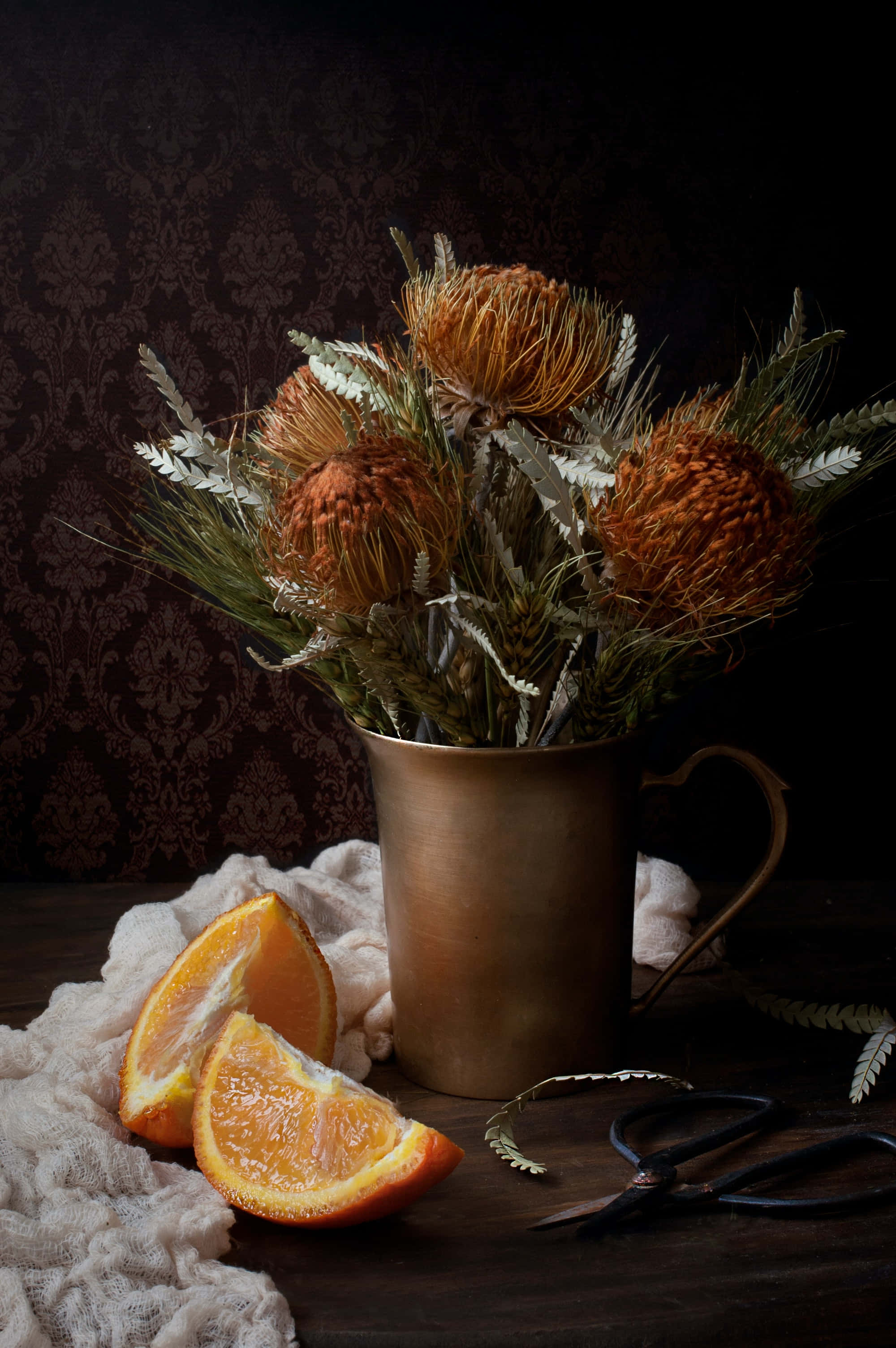 still life 桌布 壁紙 — 乾燥橙色普羅蒂亞花放在黃銅杯裡，旁邊有新鮮橙片和剪刀，放在蕾絲布上，背景是深色錦緞牆。