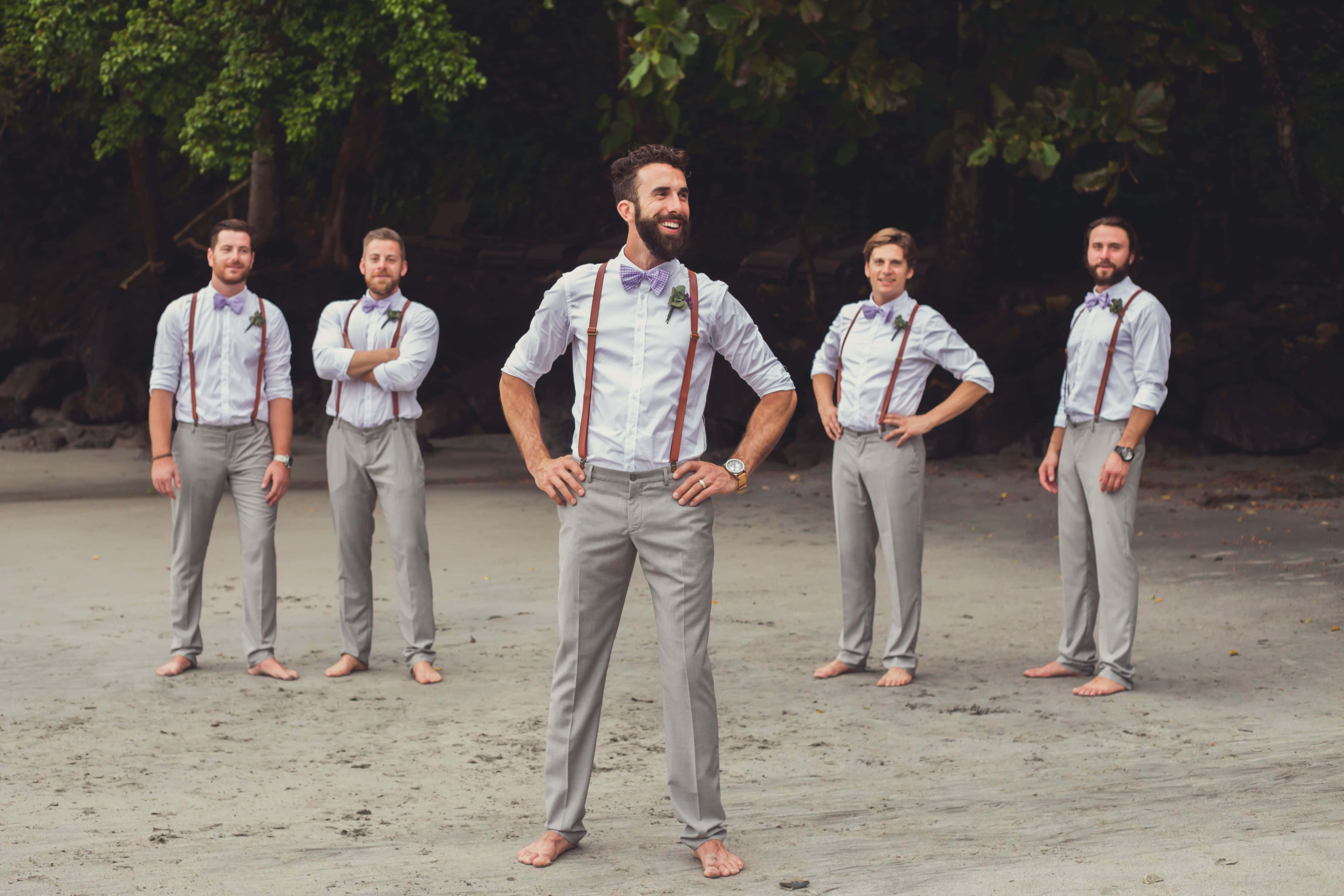 Groomsman 桌布 壁紙 — 五位伴郎穿著相配的淺色襯衫、灰色褲子和棕色吊帶，赤腳站在沙灘上，身後是茂密的綠樹。