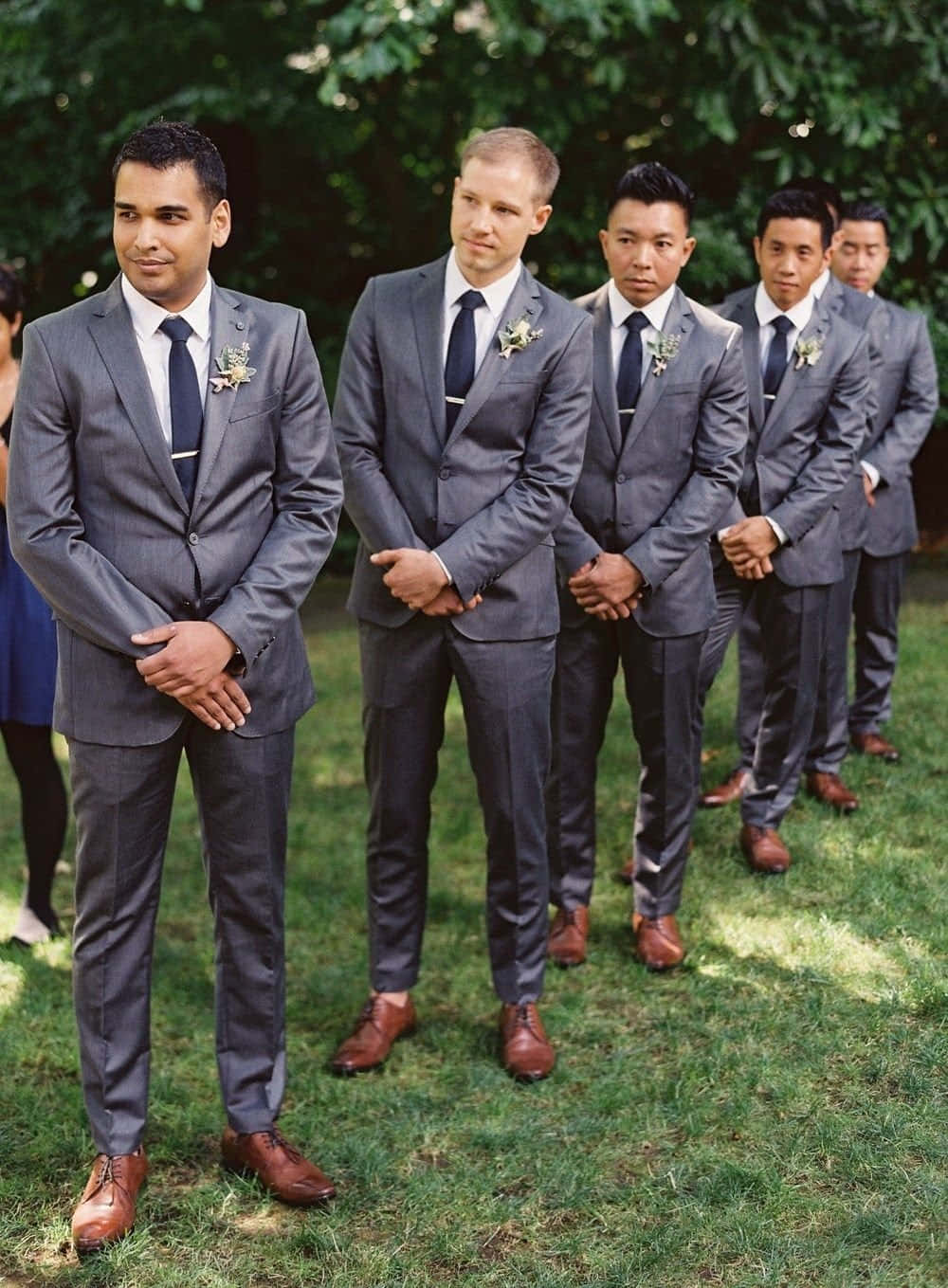 Groomsmen 桌布 壁紙 — 五位伴郎穿著相配的灰色西裝和棕色鞋子，在婚禮期間站在樹葉茂密的樹下的草地上排成一排。