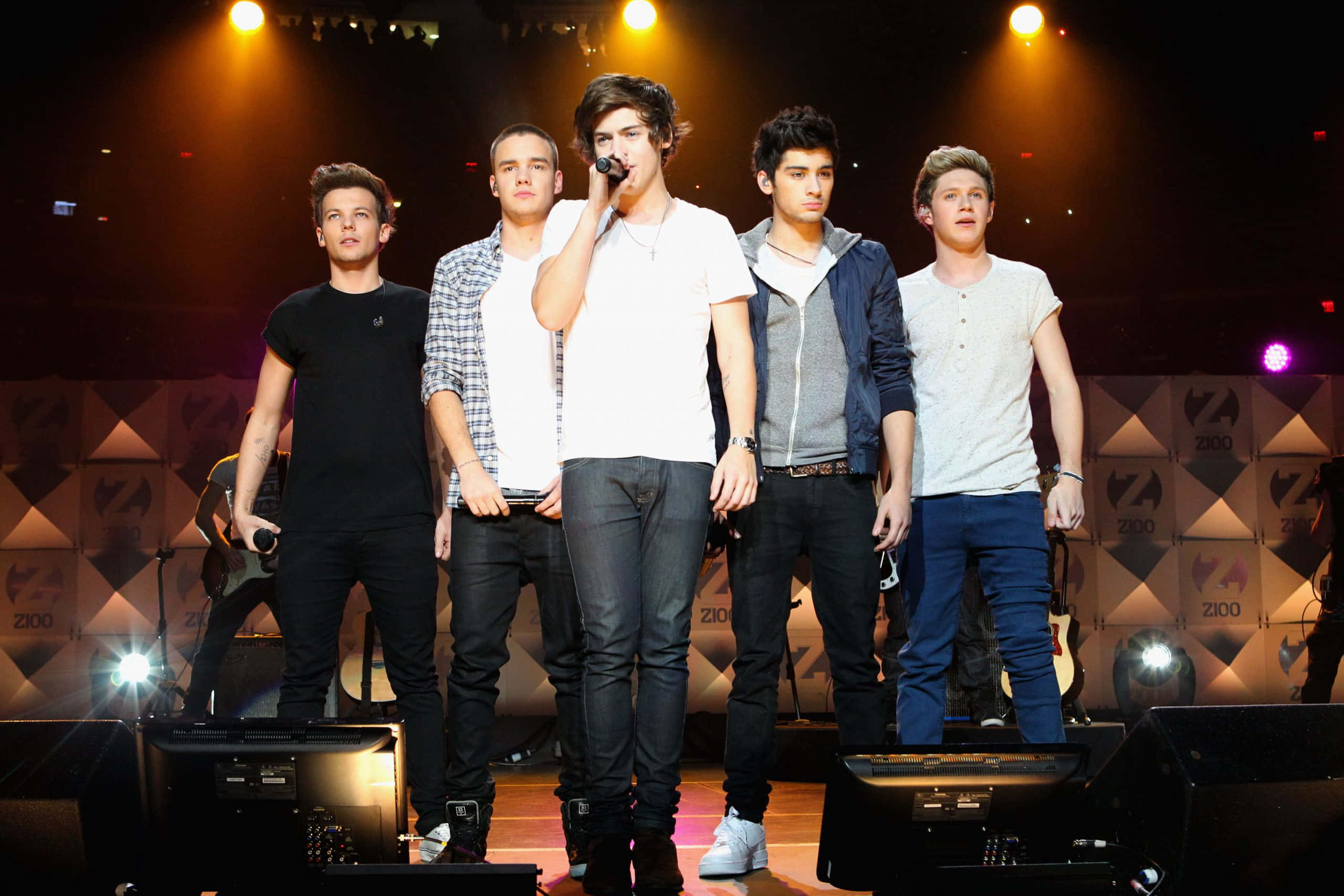 One Direction 桌布 壁紙 — 五位年輕男性樂團成員在燈光舞台上表演，其中一人對著麥克風唱歌。