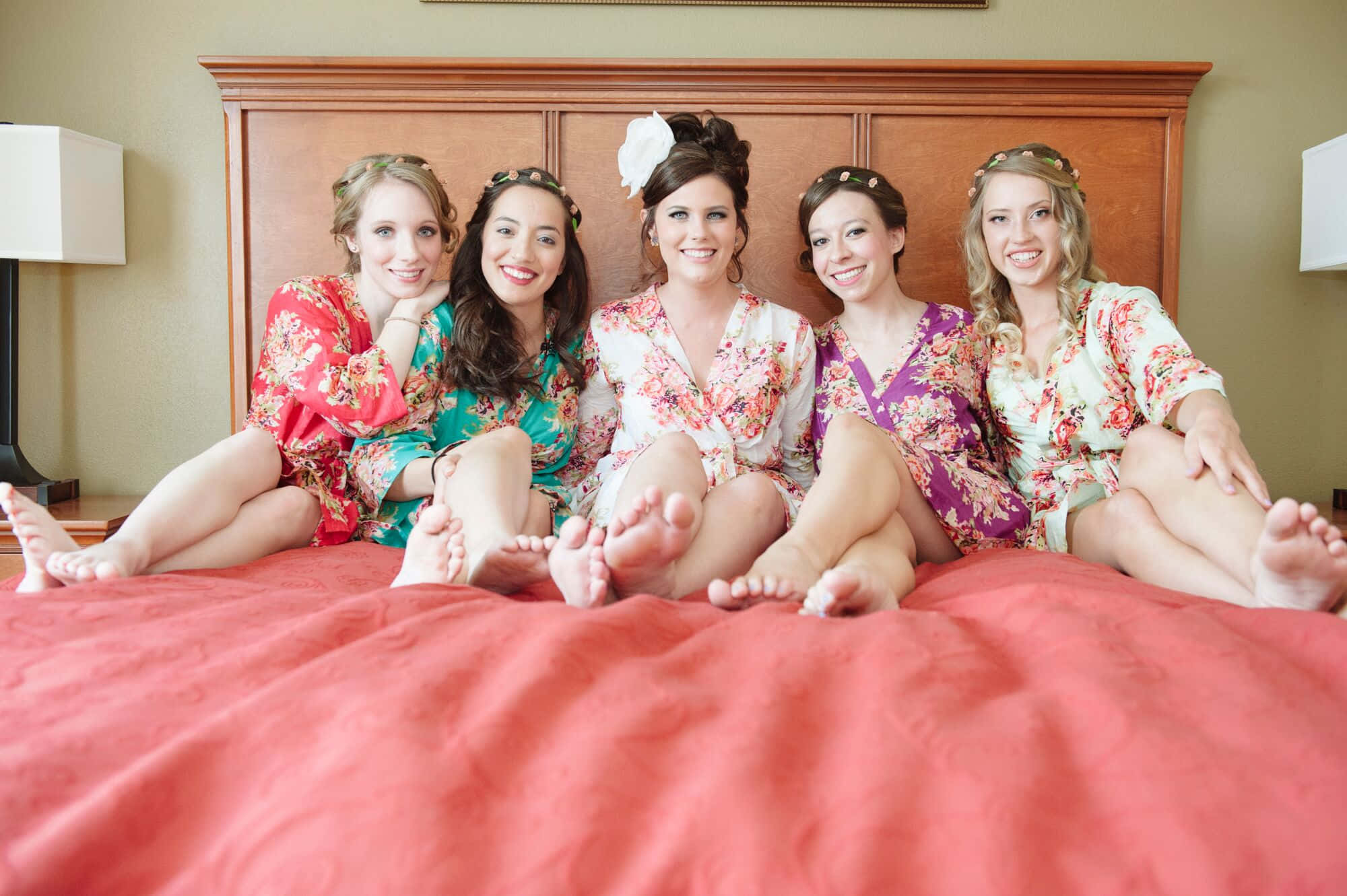 Bridesmaids 桌布 壁紙 — 五位微笑的伴娘穿著色彩繽紛的花卉睡袍，赤腳坐在珊瑚色床罩上，為婚禮前的照片愉快地擺姿勢。