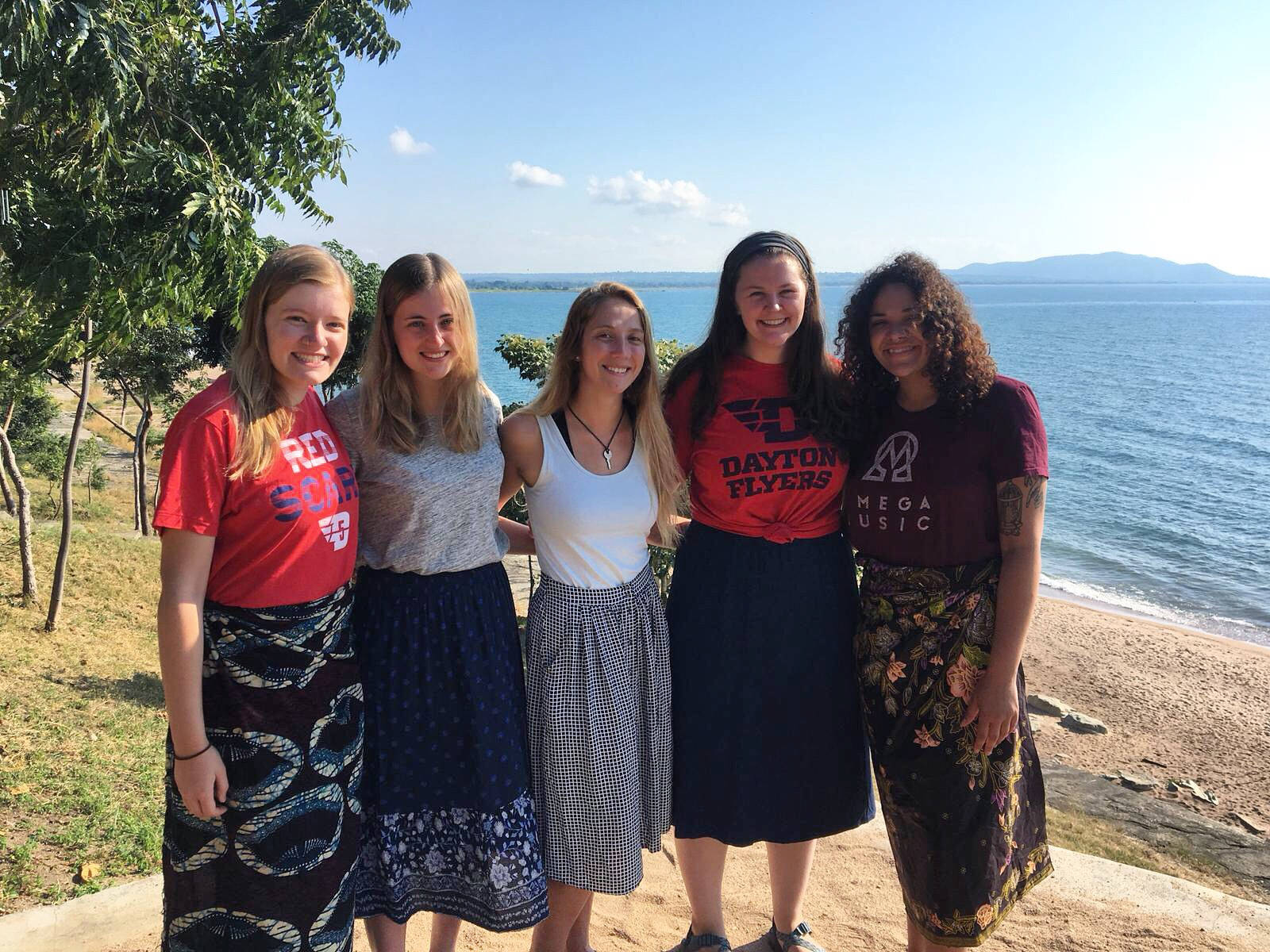 Malawi 桌布 壁紙 — 五位微笑的年輕女性穿著色彩繽紛的裙子，一起站在陽光明媚的海邊懸崖上，俯瞰平靜的藍色大海。