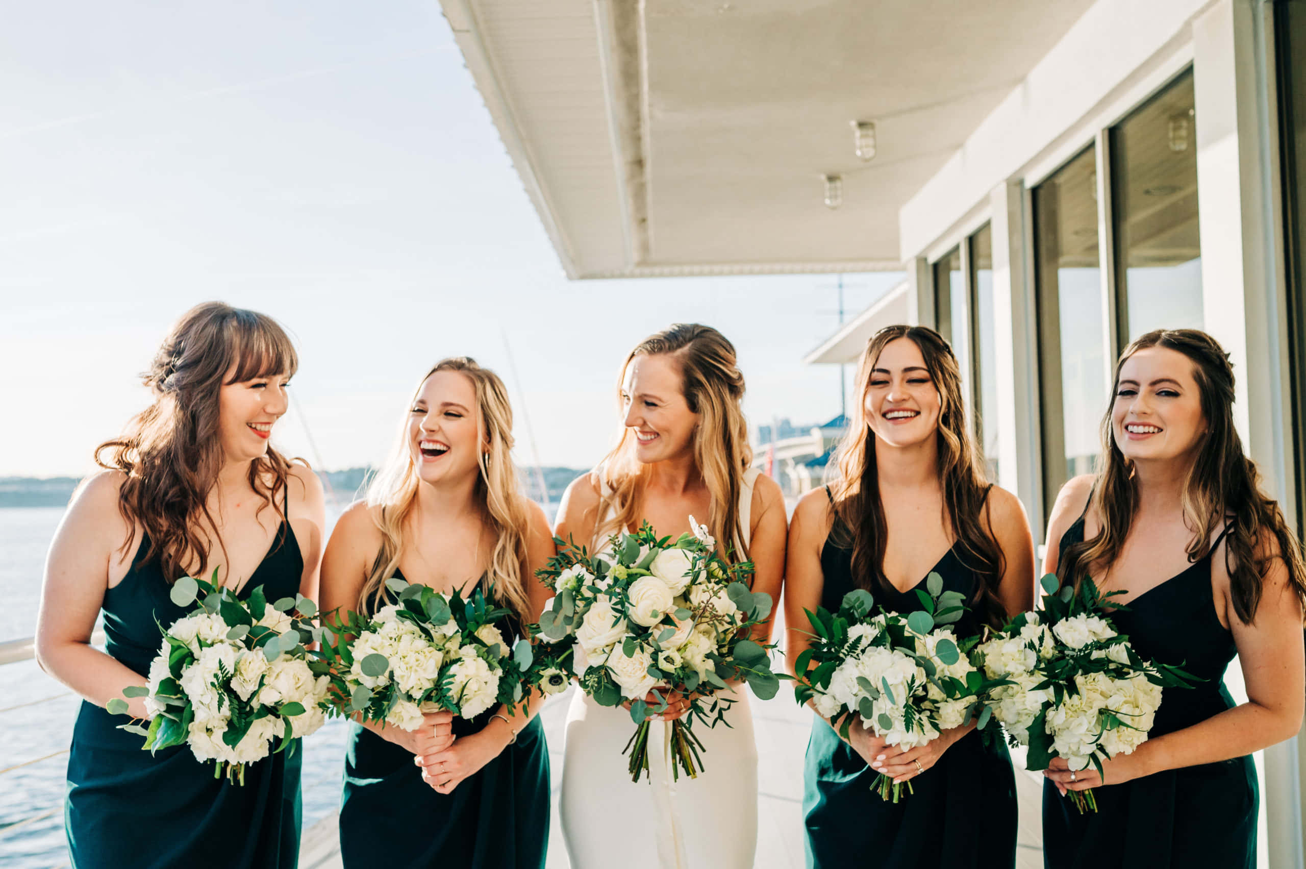 Bridal 桌布 壁紙 — 五位快樂的伴娘穿著深綠色連衣裙，在陽光明媚的海濱陽台上一起笑著，手持白色花束。