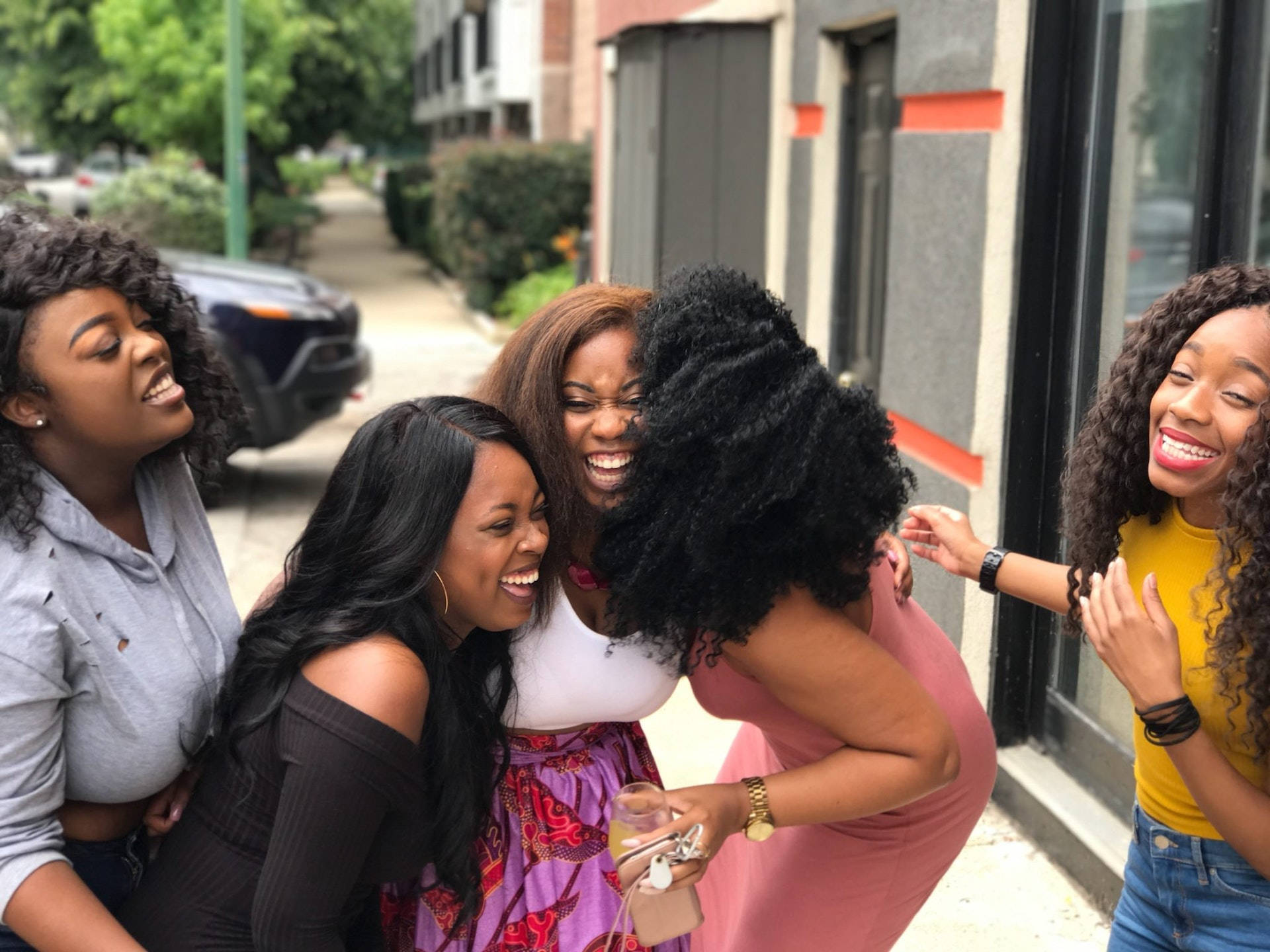 Sexy Black Women 桌布 壁紙 — 五位快樂的女性在陽光明媚的城市人行道上，被綠植和建築環繞，大笑並緊緊擁抱。