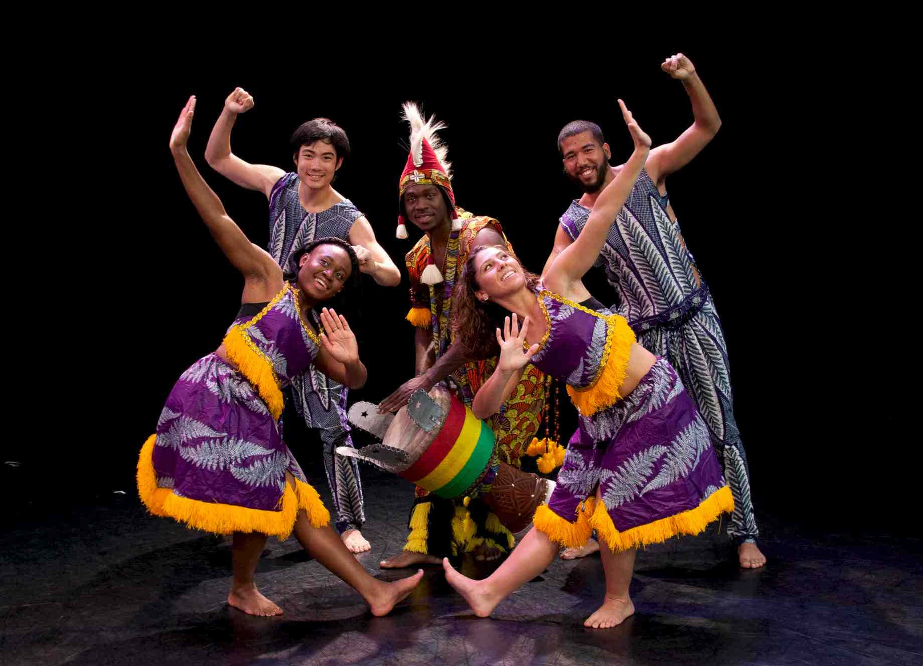 African Culture 桌布 壁紙 — 五位快樂的舞者穿著鮮豔的紫色和黃色服裝，在一個帶有彩色鼓的黑暗舞台上擺出動態姿勢。