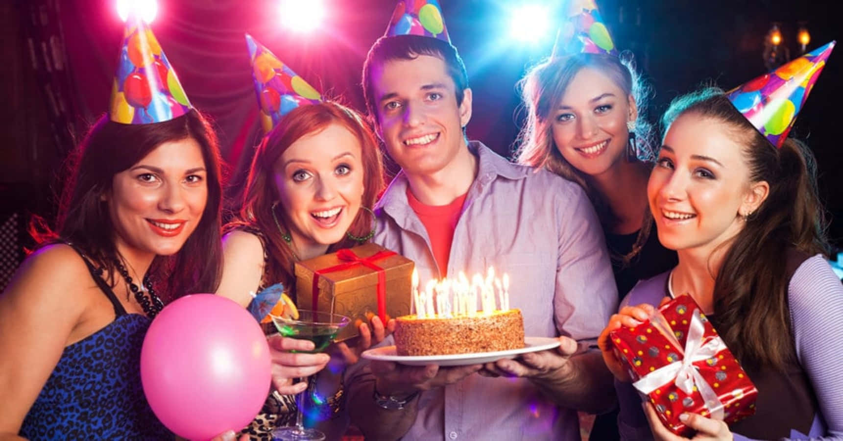 Birthday Celebration 桌布 壁紙 — 五位戴著派對帽、面帶微笑的朋友，在色彩繽紛的舞台燈光下，圍繞著一個插著點燃蠟燭的生日蛋糕。