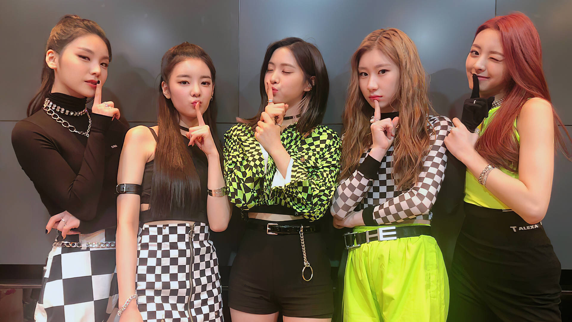 ITZY 桌布 壁紙 — 五位時尚女性穿著大膽的格紋和螢光服裝，在黑暗的攝影棚牆前擺出手指放在嘴唇上的姿勢。