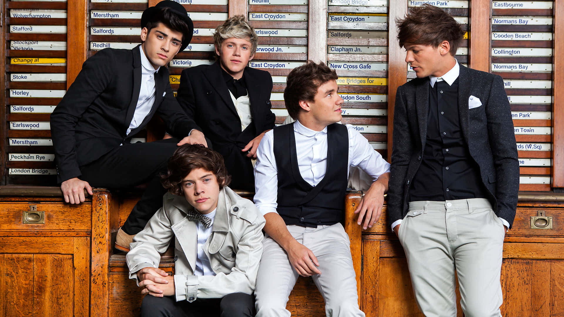One Direction 桌布 壁紙 — 五位時尚年輕男士身穿正裝，自信地坐在復古木長椅上，背景是舊火車站標誌。