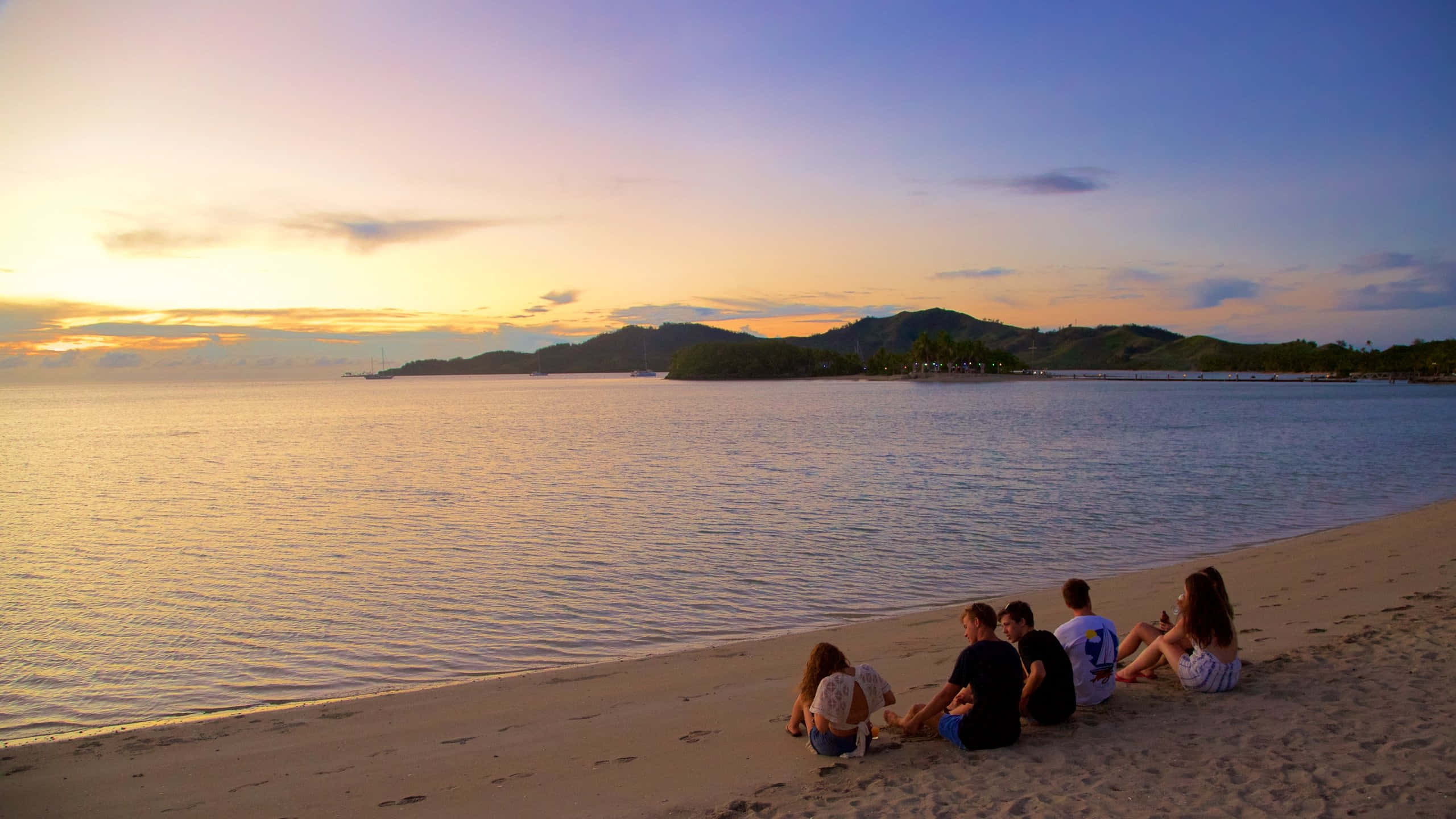 Fiji Beach 桌布 壁紙 — 五位朋友坐在沙灘上，欣賞日落時金色天空映照在平靜海面上，遠方有綠色山丘。
