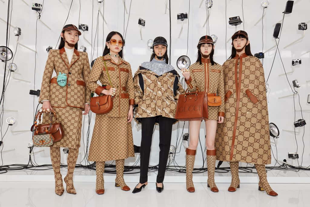 Gucci 桌布 壁紙 — 五位模特兒穿著Gucci標誌性的字母組合服裝，自信地擺姿勢，背景是裝飾著相機和燈光的白色牆壁。