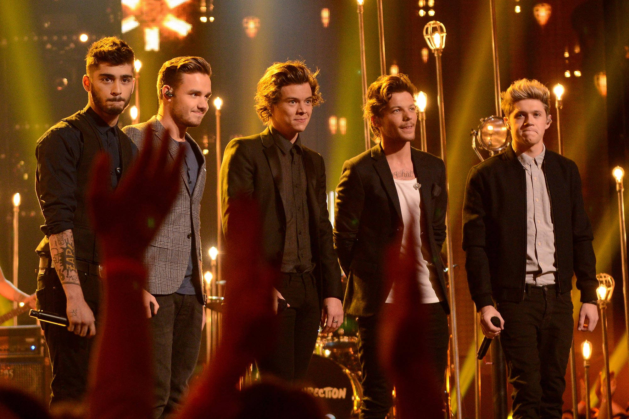 One Direction Aesthetic 桌布 壁紙 — 五位男性樂隊成員自信地站在一個溫暖照明的舞台上，帶有發光的垂直燈光和前景中舉起的觀眾手。
