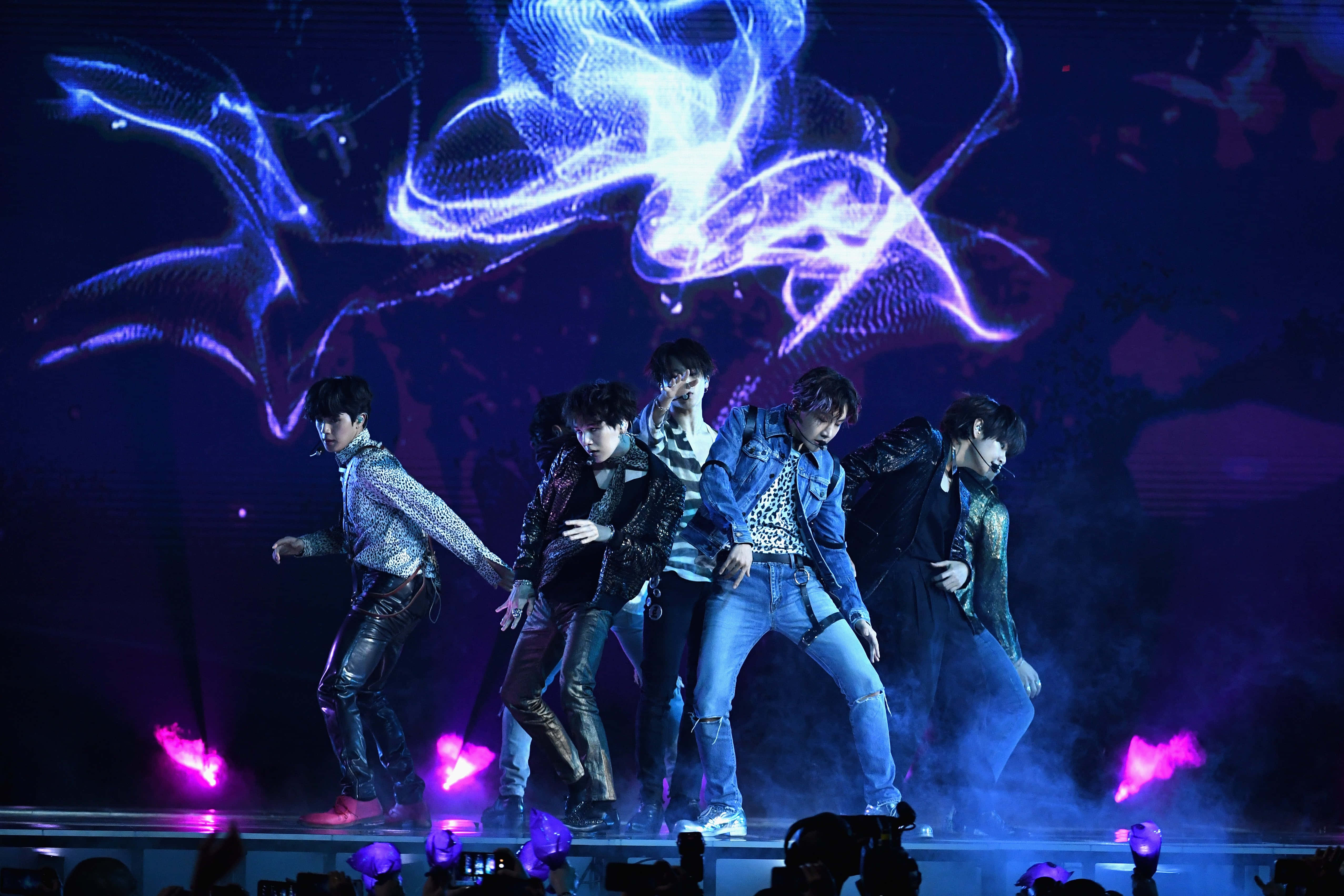 Bts Performance 桌布 壁紙 — 五位男性表演者在閃爍的藍色和紫色舞台燈光下擺出動感舞蹈姿勢，身後有旋轉的數位投影。