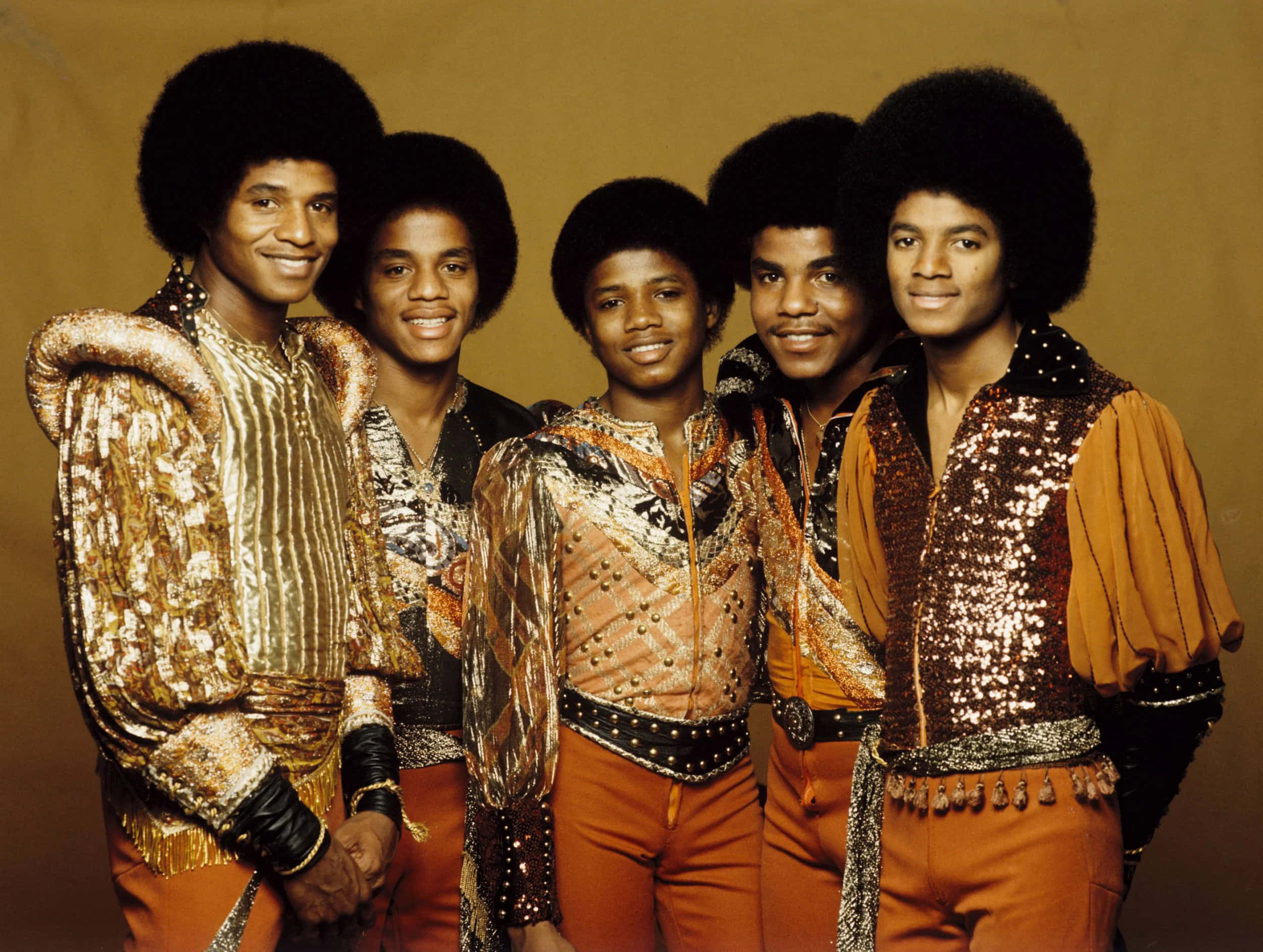 Young Michael Jackson 桌布 壁紙 — 五位留著爆炸頭的年輕男士，在溫暖的棕色背景前穿著閃閃發光的金色和橙色亮片服裝。
