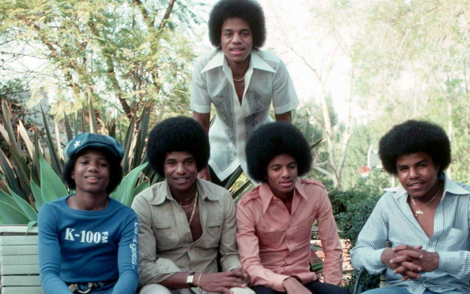 Jackson 5 桌布 壁紙 — 五位留著非洲式髮型、身穿1970年代服裝的男士在戶外綠意盎然和植物高聳的環境中微笑。