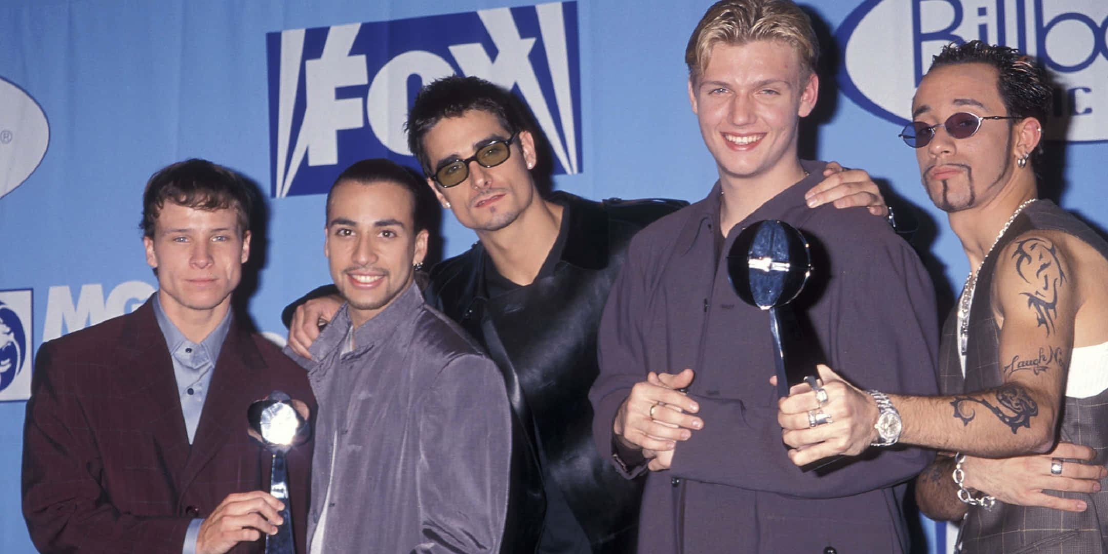 Backstreet Boys 桌布 壁紙 — 五位身穿1990年代時尚的男士，在藍色Fox和Billboard背景前手持獎盃擺姿勢。