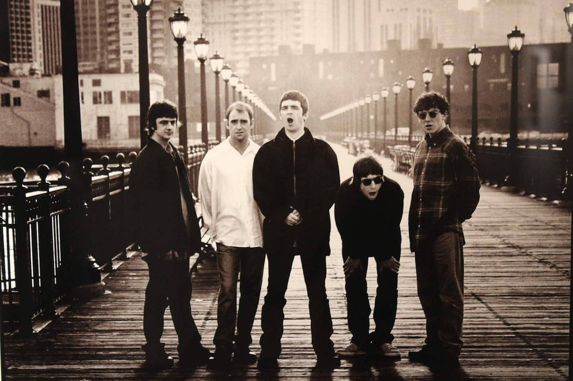 綠洲 桌布 壁紙 — 五位身穿復古街頭服飾的男性在木製碼頭上戲劇性地擺姿勢，背景是城市天際線和路燈，色調為懷舊棕色。