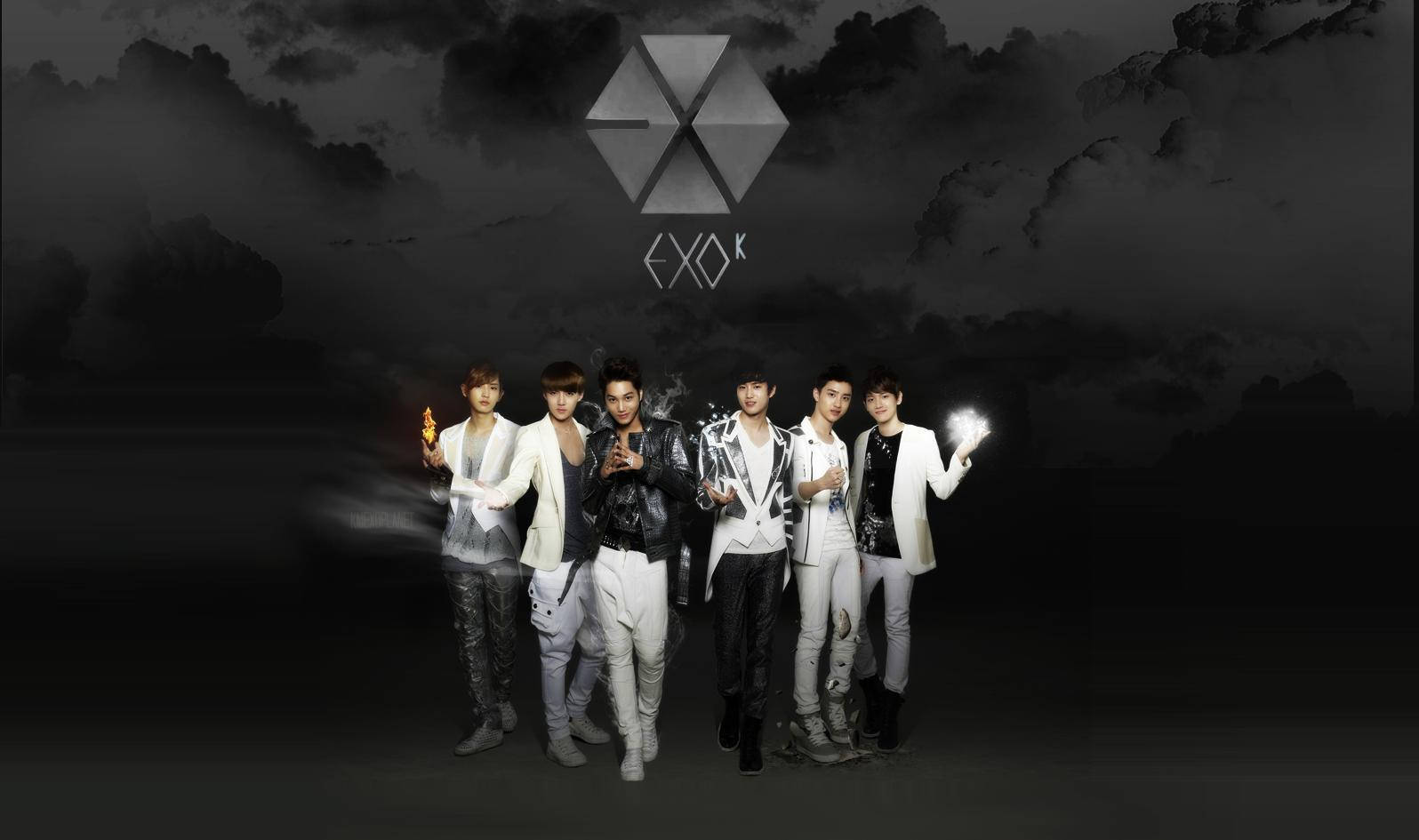 K-pop 桌布 壁紙 — 五位身穿時尚黑白服裝的男士站在陰暗多雲的天空下，手持發光球體，上方有一個幾何標誌。