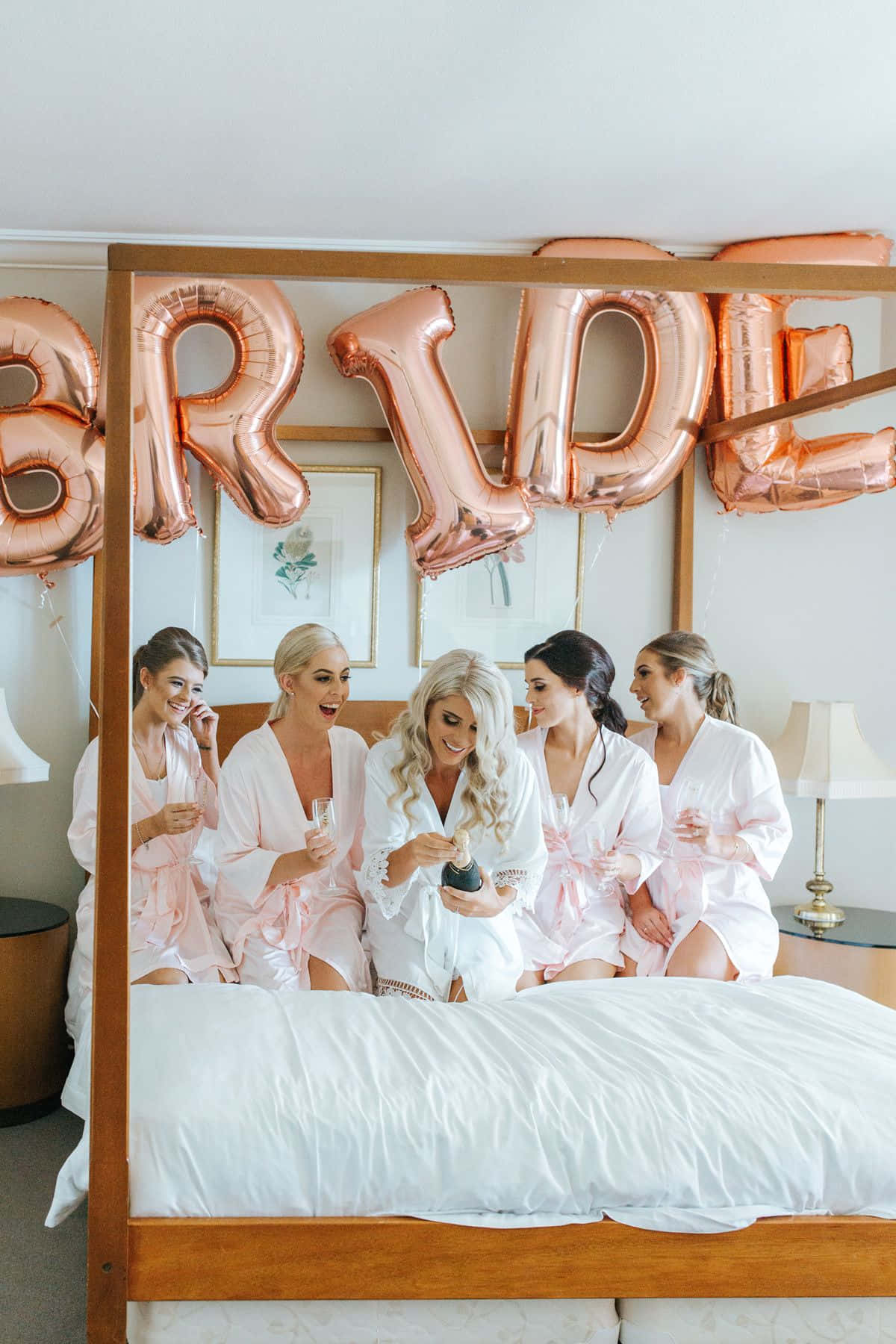 Bride 桌布 壁紙 — 五位身穿粉紅色長袍的女性在床上開懷大笑，新娘在玫瑰金色「BRIDE」氣球下打開香檳。