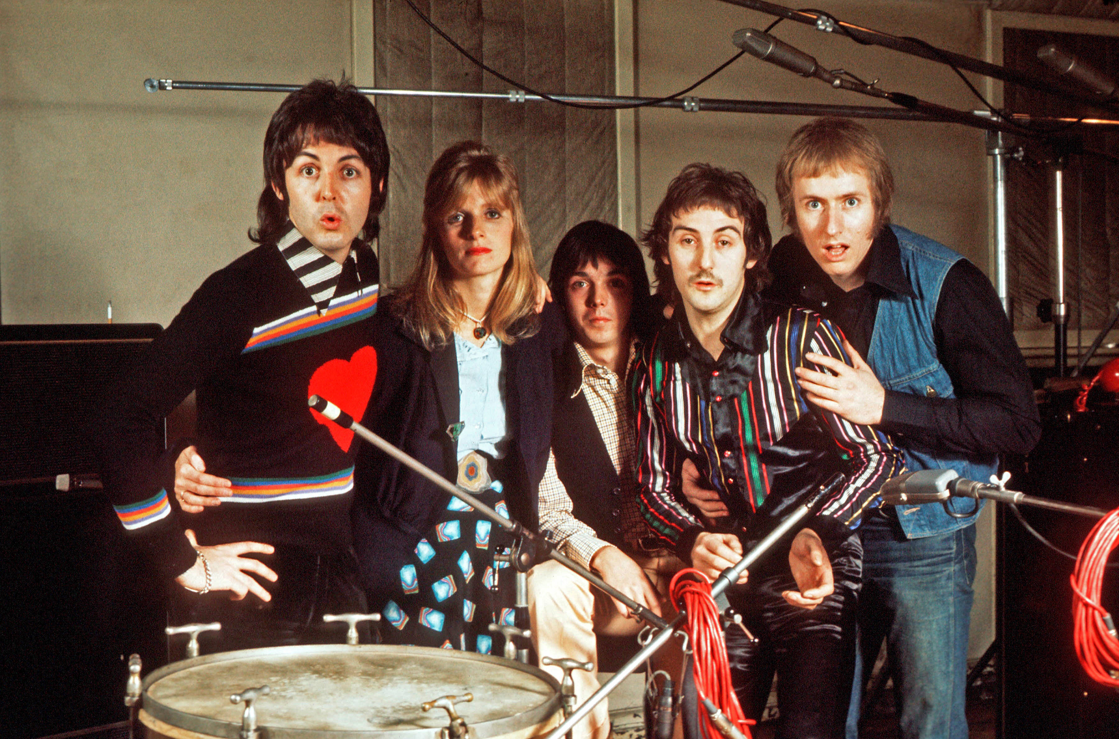 paul mccartney 桌布 壁紙 — 五位身穿色彩繽紛70年代服裝的音樂家在錄音室內圍繞麥克風和鼓擺出戲劇性的姿勢。