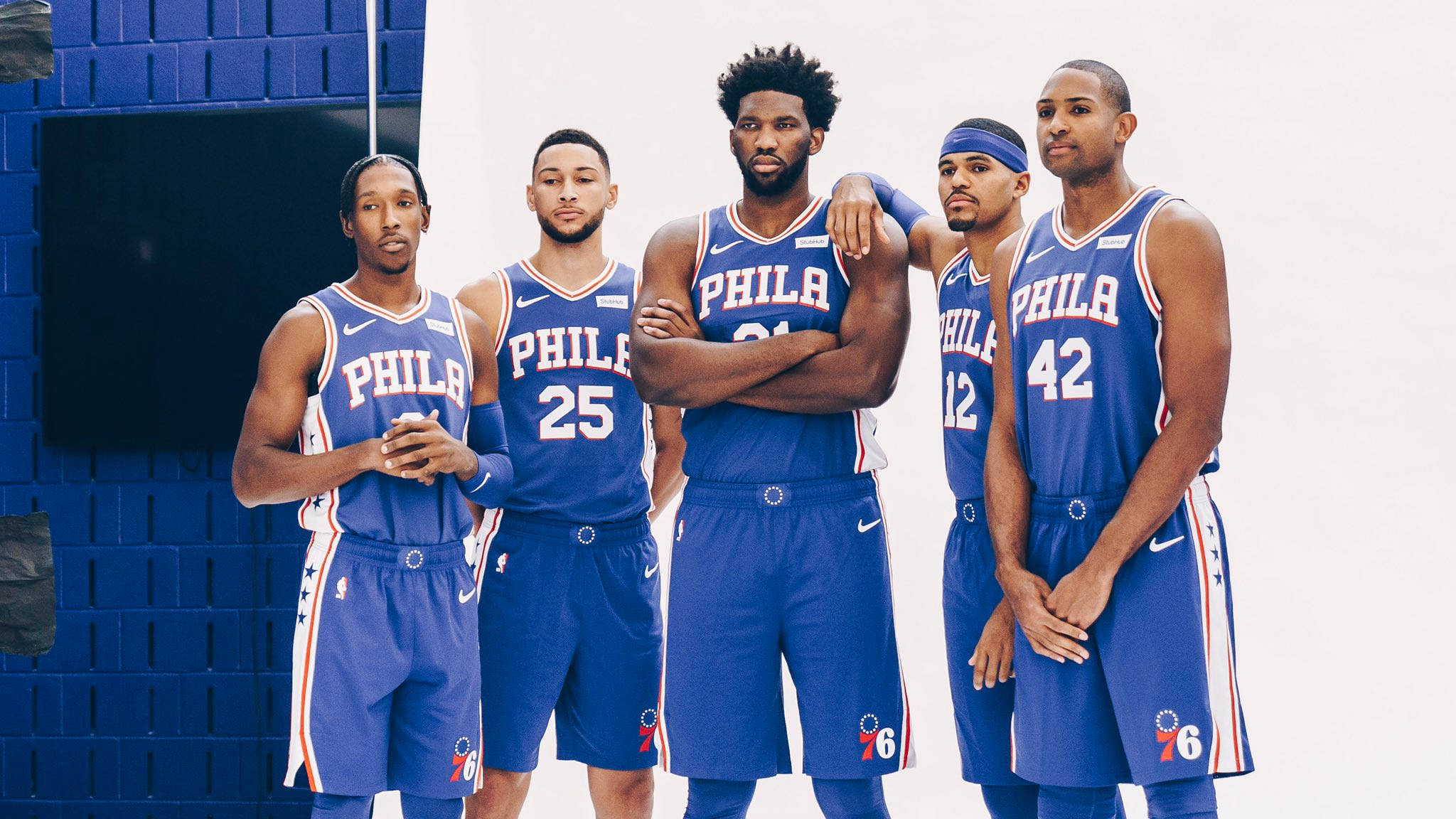 76ers 桌布 壁紙 — 五位身穿藍色球衣的費城76人籃球運動員在藍色磚牆和白色背景前自信地擺姿勢。