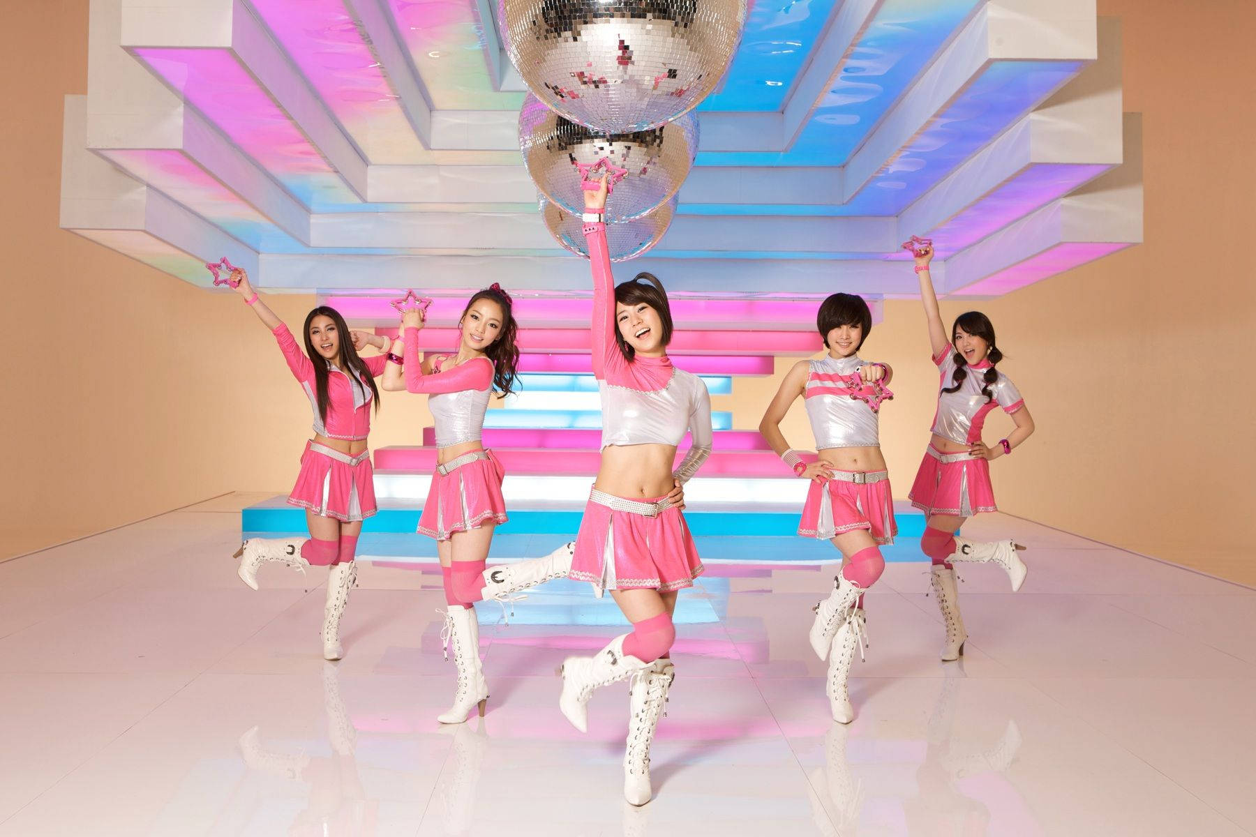 K-pop 桌布 壁紙 — 五位面帶微笑的舞者穿著粉紅色和銀色服裝，在未來主義的霓虹燈工作室中，在迪斯科球下充滿活力地擺姿勢。