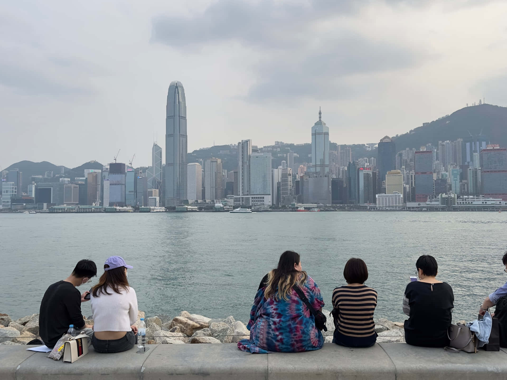 香港 桌布 壁紙 — 五個人坐在石壆上，俯瞰陰天下香港標誌性的天際線和港口。