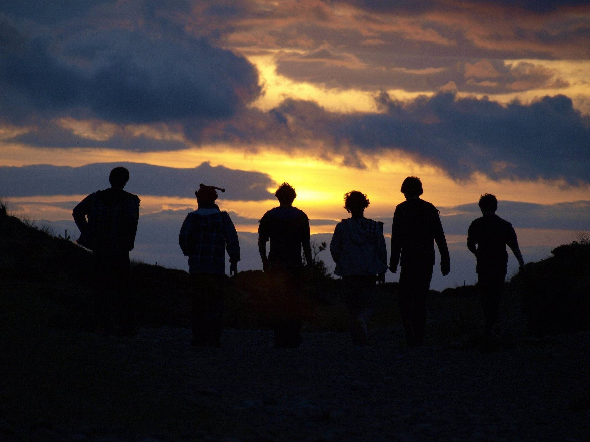 Silhouette 桌布 壁紙 — 五個剪影人物沿著山坡行走，夕陽西下，上方有戲劇性的橙色和紫色雲彩。